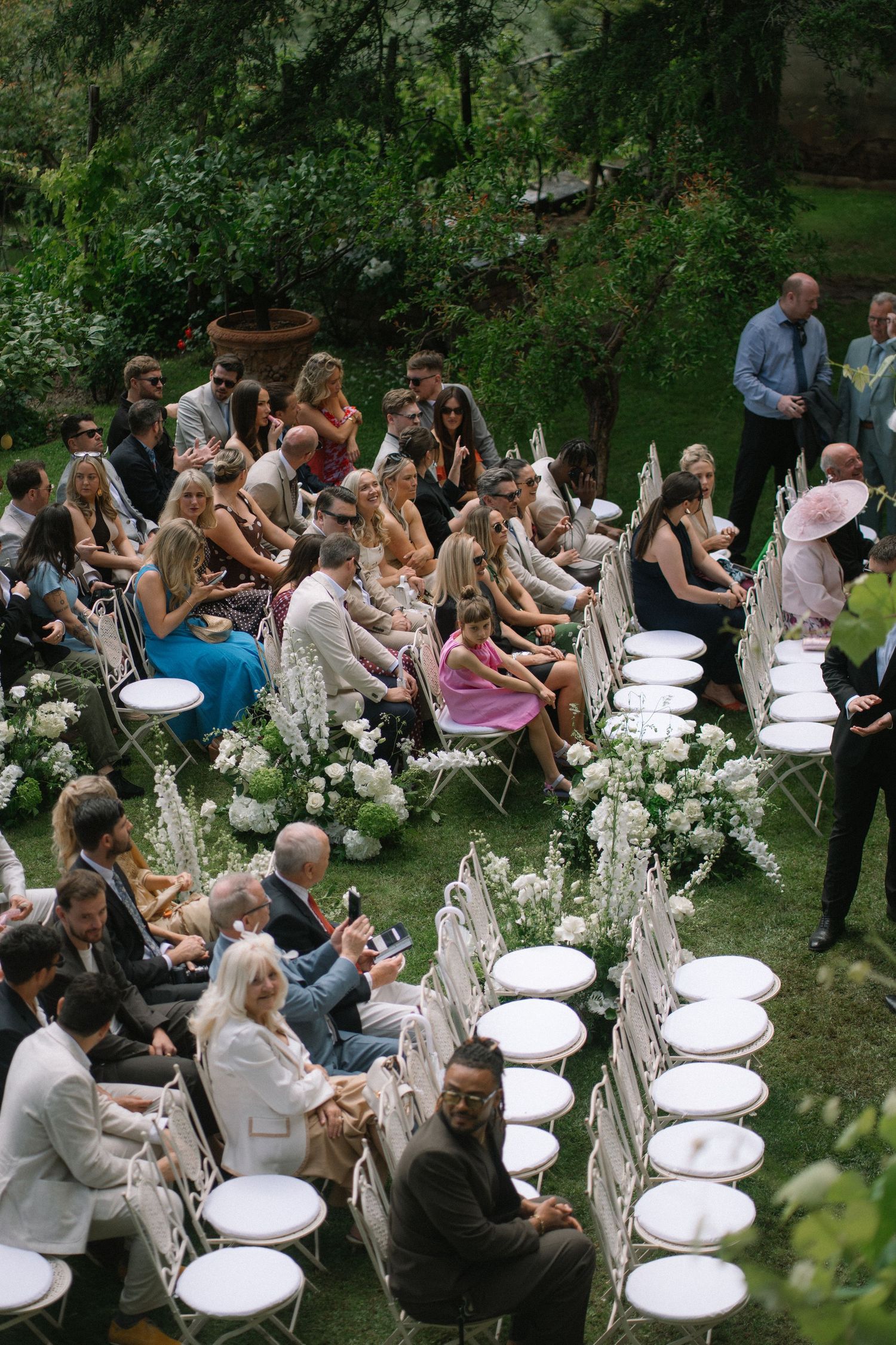 Aerial view of wedding guests seated in white chairs at an outdoor garden ceremony with white floral arrangements.