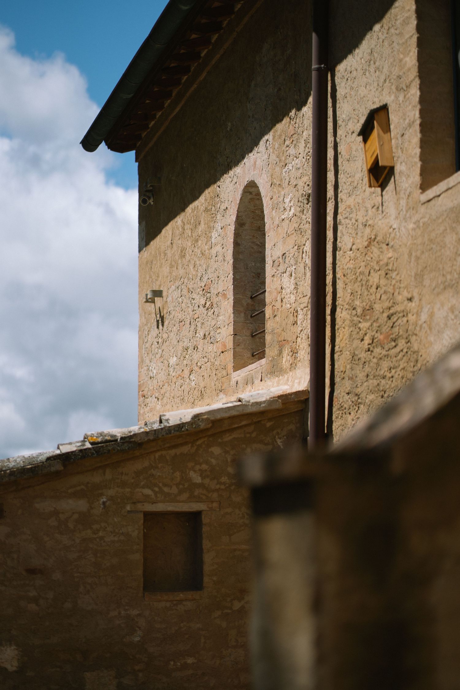 Ancient stone building with arched window and rustic facade under a partly cloudy blue sky in Tuscany.