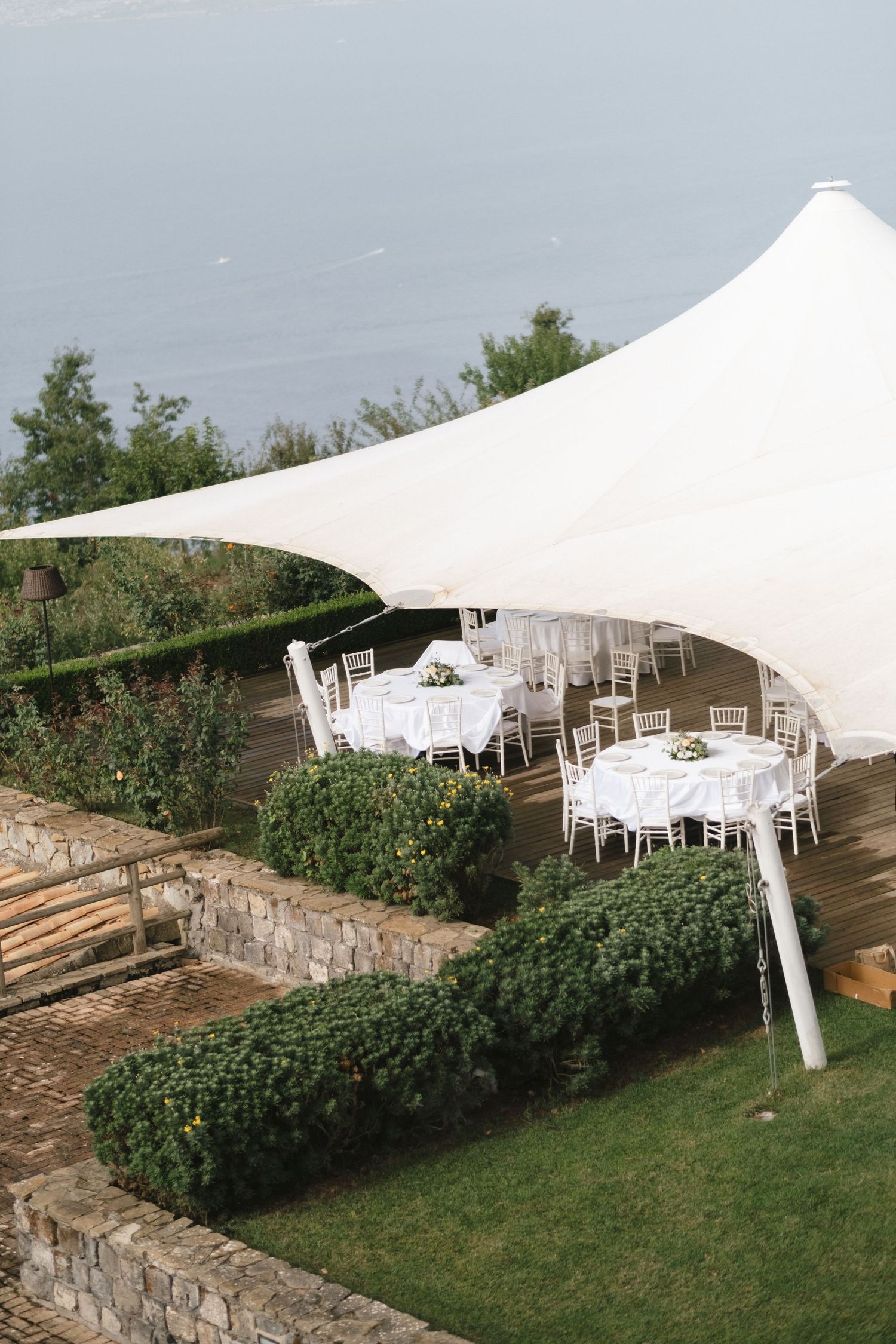 Elegant outdoor wedding reception under a white tensile canopy with round tables and chairs overlooking a scenic garden.