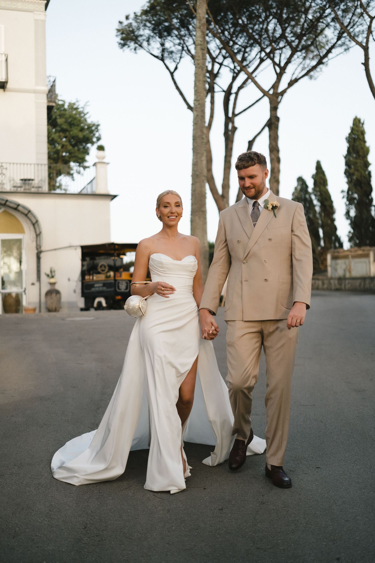 Bride and groom walk hand in hand, she in strapless white gown, he in beige suit, at Italian venue.