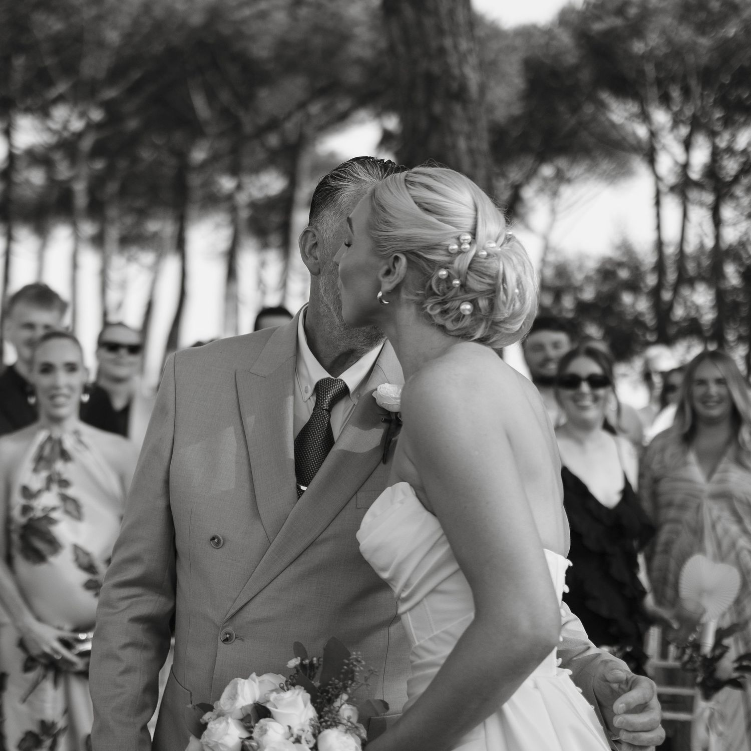 Black and white photo of a bride and groom sharing a kiss at their outdoor wedding ceremony with guests.