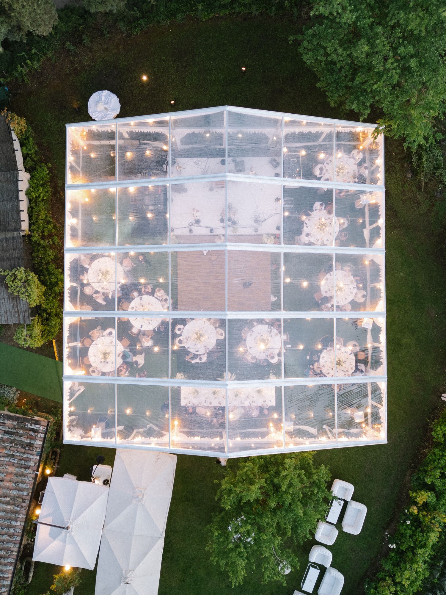 Aerial view of a clear wedding reception tent with round tables and floral centerpieces surrounded by lush greenery.