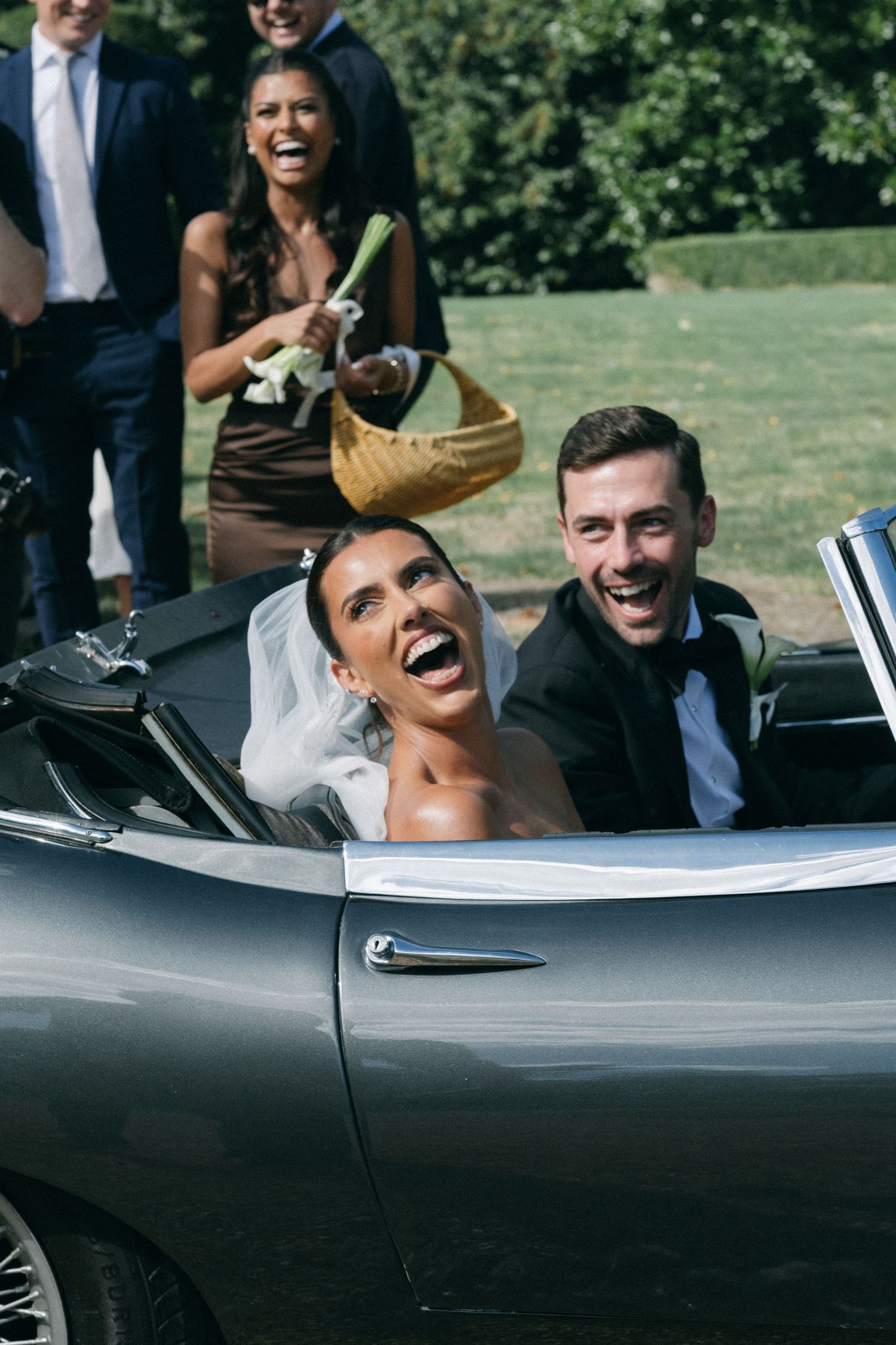 Joyful bride and groom laughing in a silver convertible classic car, with wedding guests celebrating behind them.