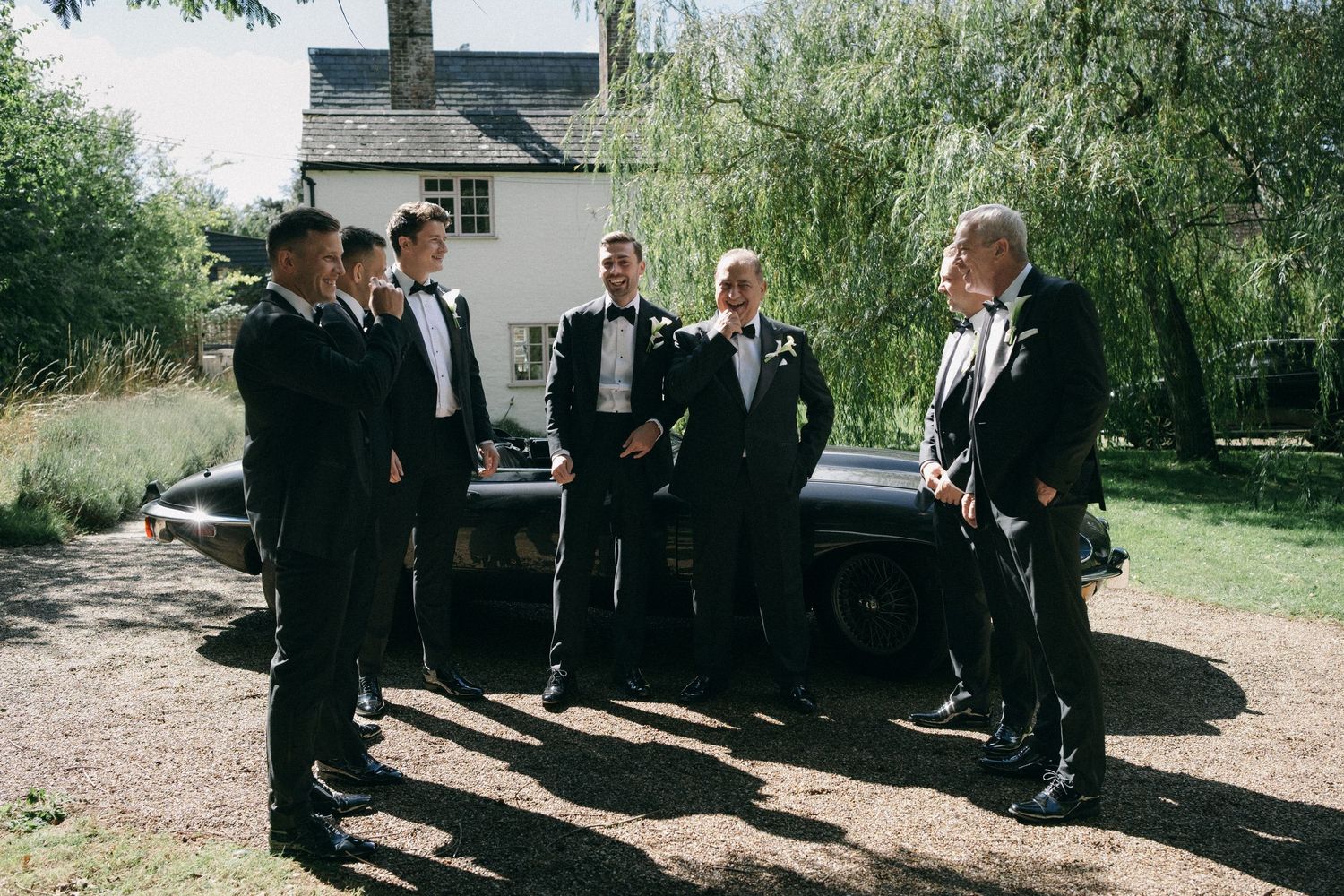 Groomsmen in black tuxedos standing beside a vintage black car outdoors near a white house on a sunny wedding day.