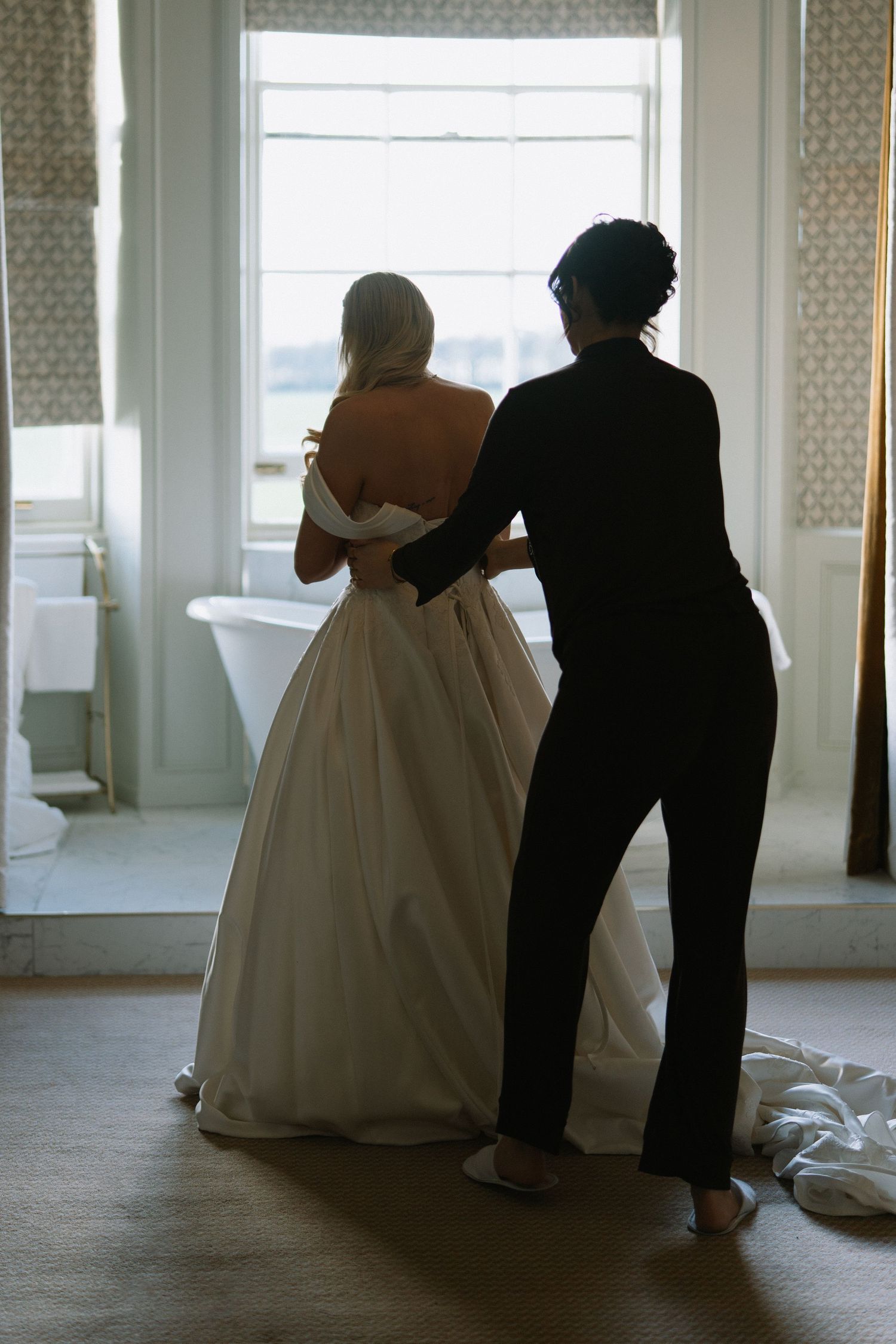 A bride in a white ball gown being helped by a woman in black near a bright window.