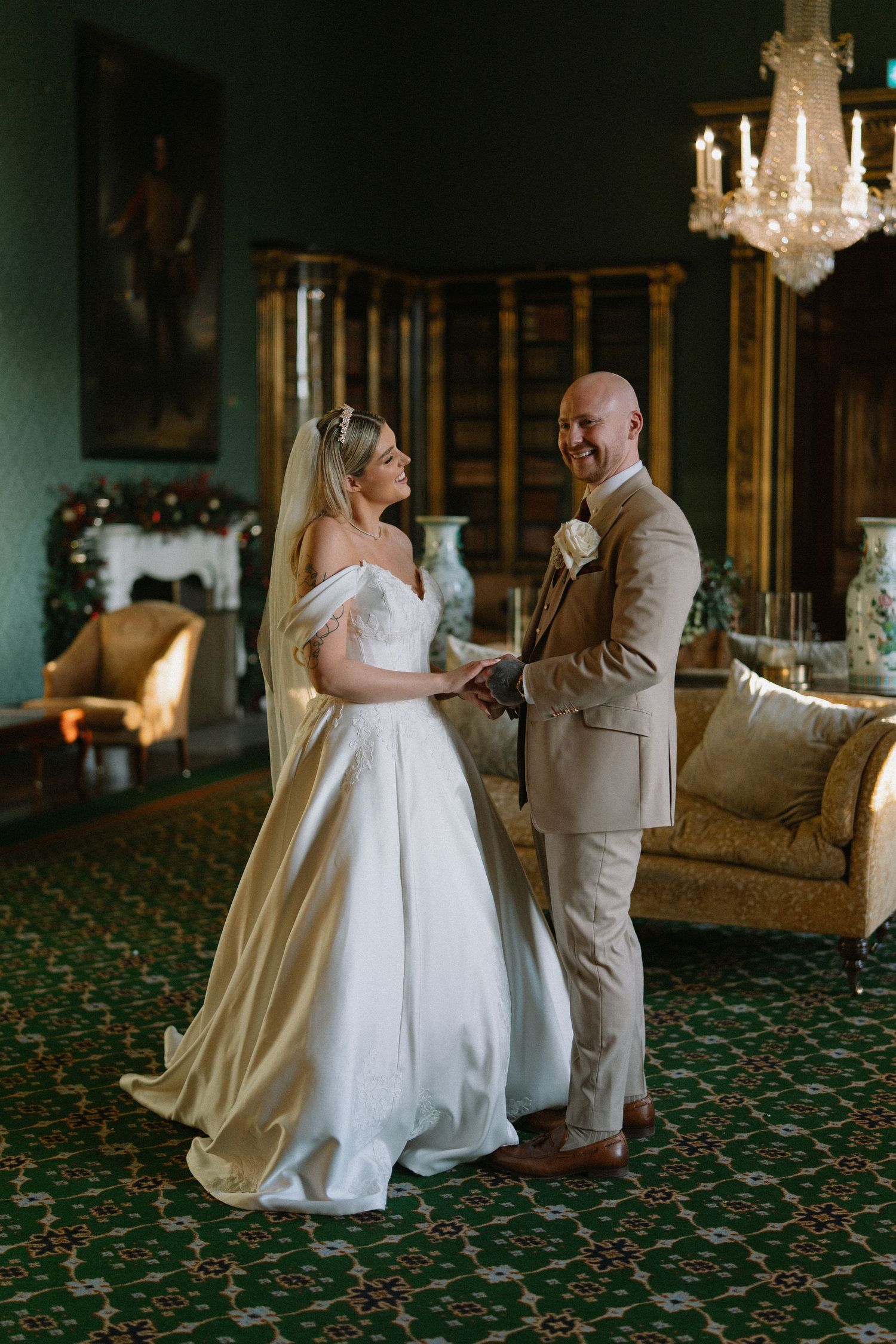 Bride and groom holding hands smiling in elegant green room with chandelier and fireplace.