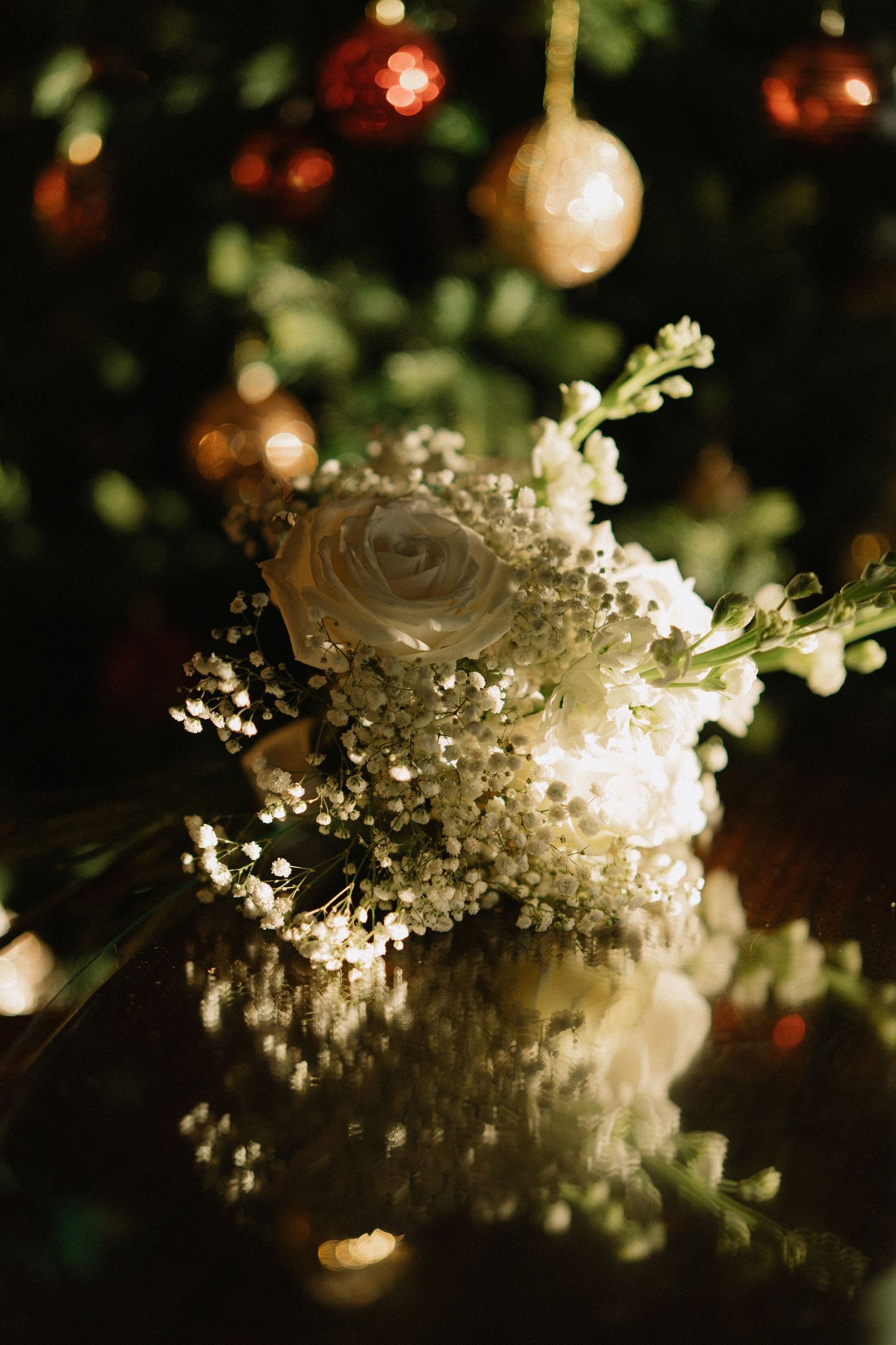 White rose and baby's breath bouquet reflecting on dark surface with Christmas ornaments in background.