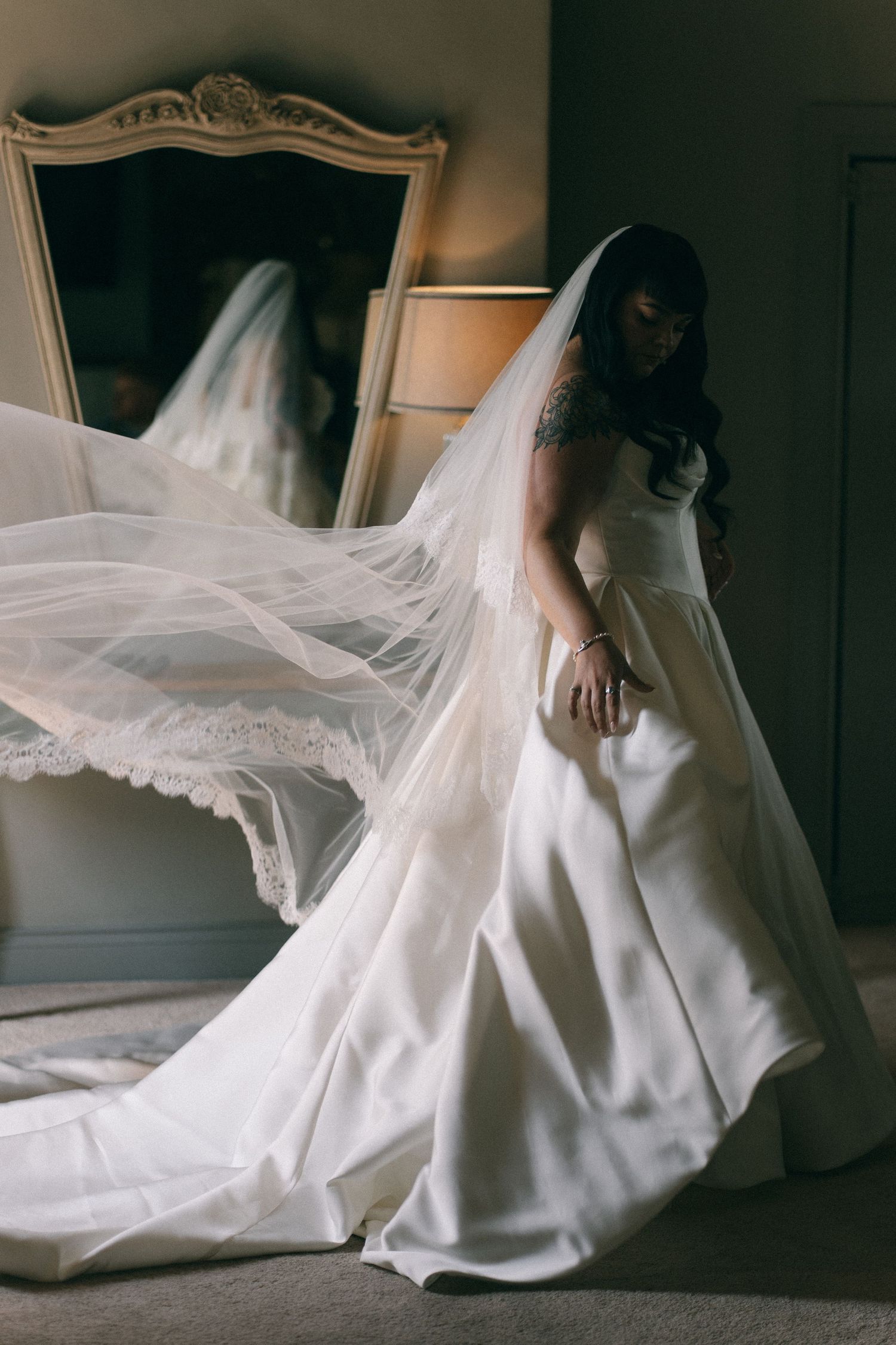 Bride in a flowing white wedding gown with lace-trimmed veil billowing gracefully in a softly lit room.