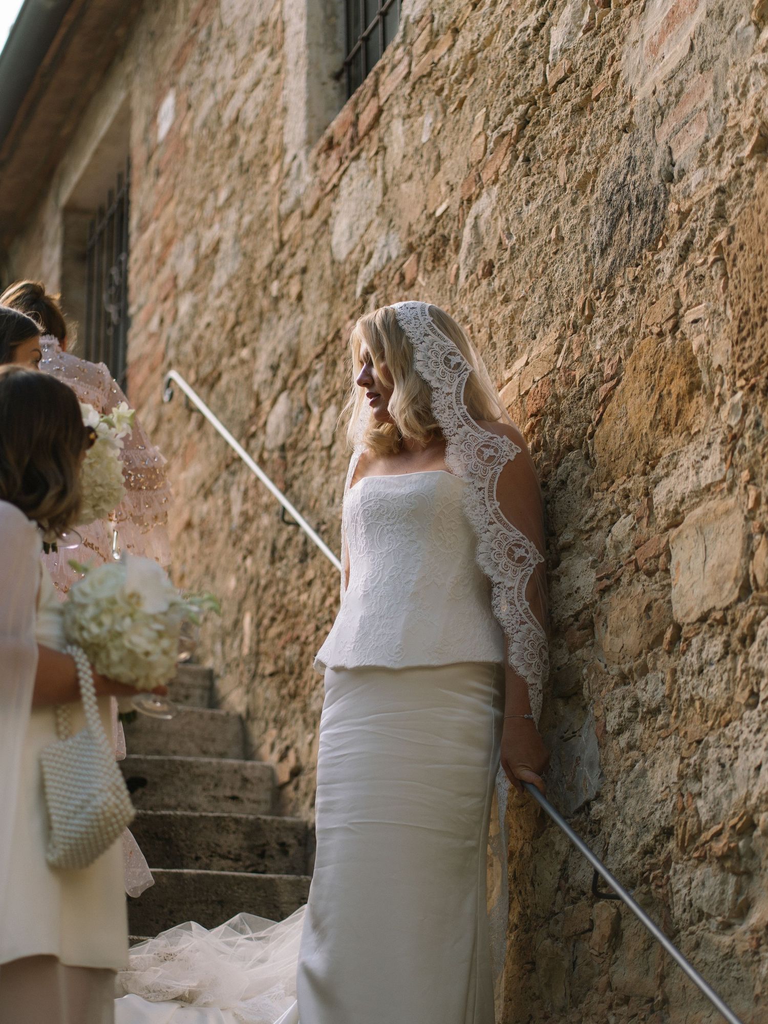 Bride in white lace peplum gown and mantilla veil leaning against rustic stone wall with bridesmaids.