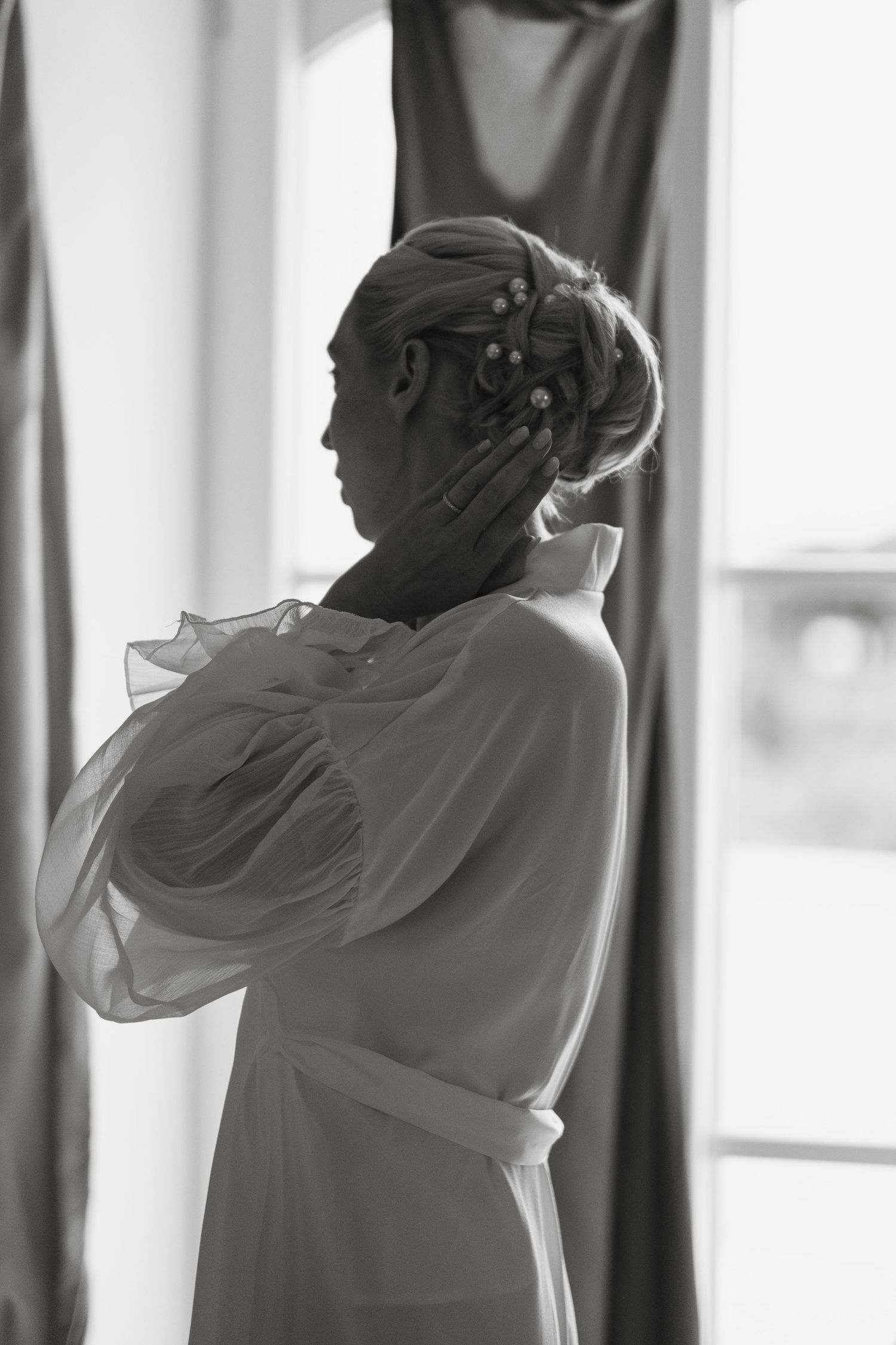 Black and white photo of a bride in a sheer robe touching her face, showcasing an elegant updo with pearl hair accessories.