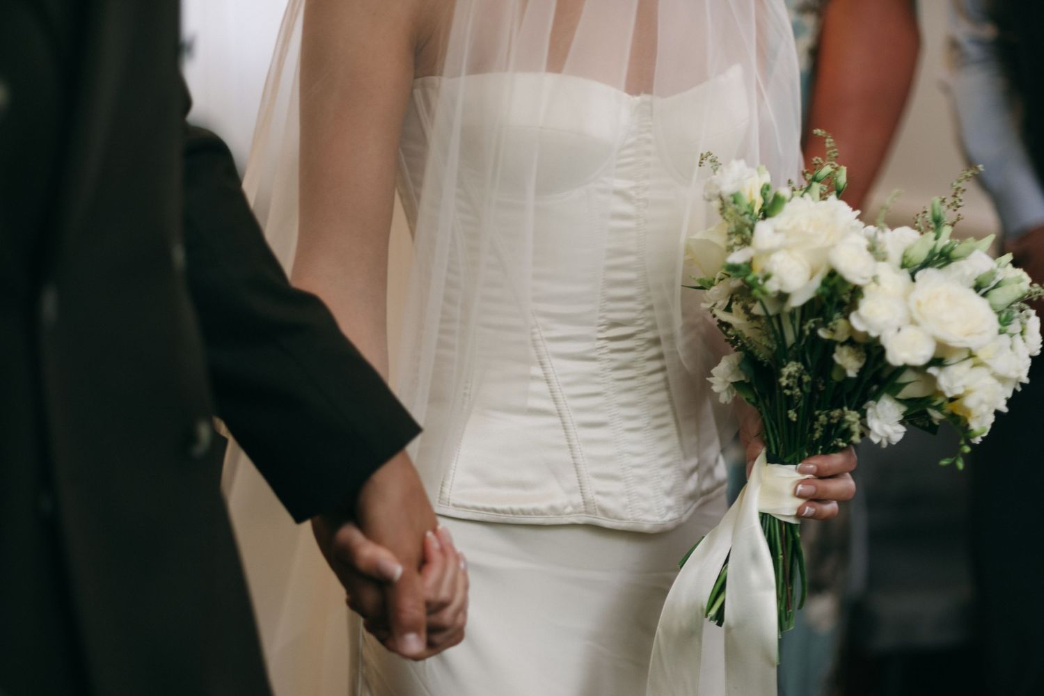 Bride in white strapless gown holding bouquet of white roses, holding hands with groom in black suit.