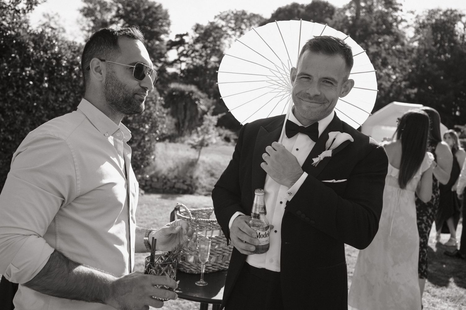 Black and white photo of two men in formal attire holding drinks and a parasol at an outdoor wedding reception.