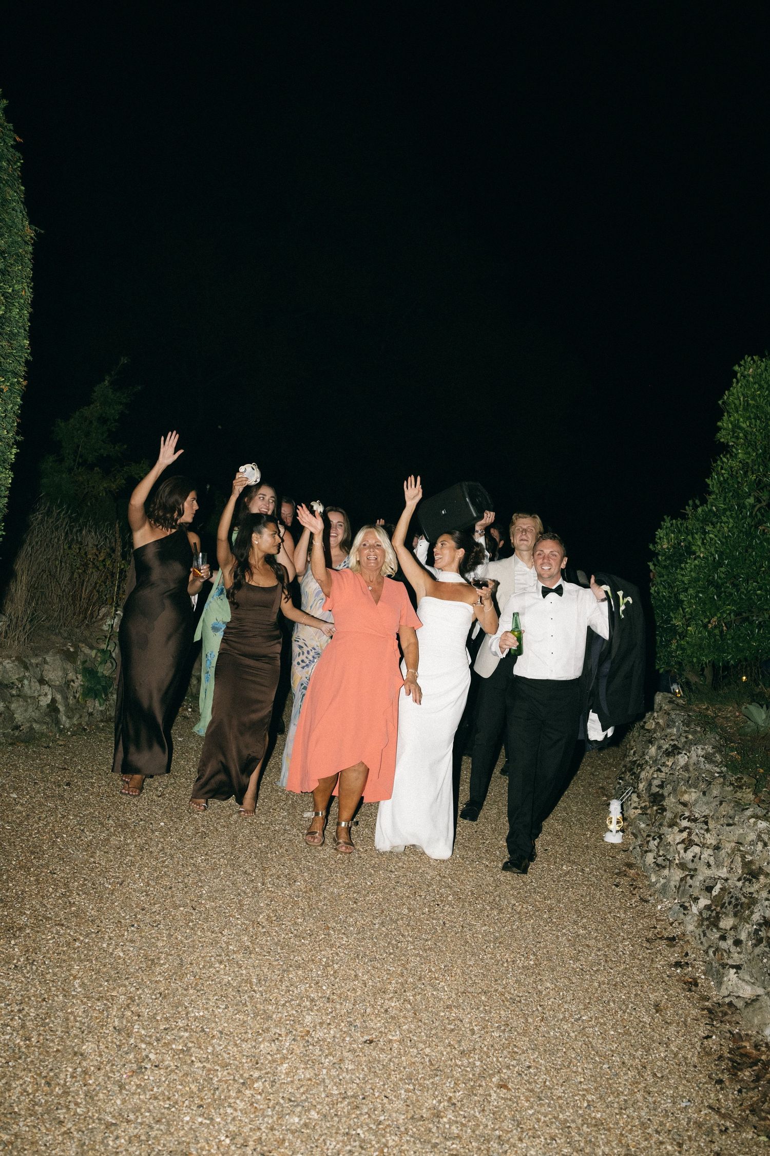 Joyful wedding party celebrating outdoors at night, bride in white gown with groom in tuxedo surrounded by guests.