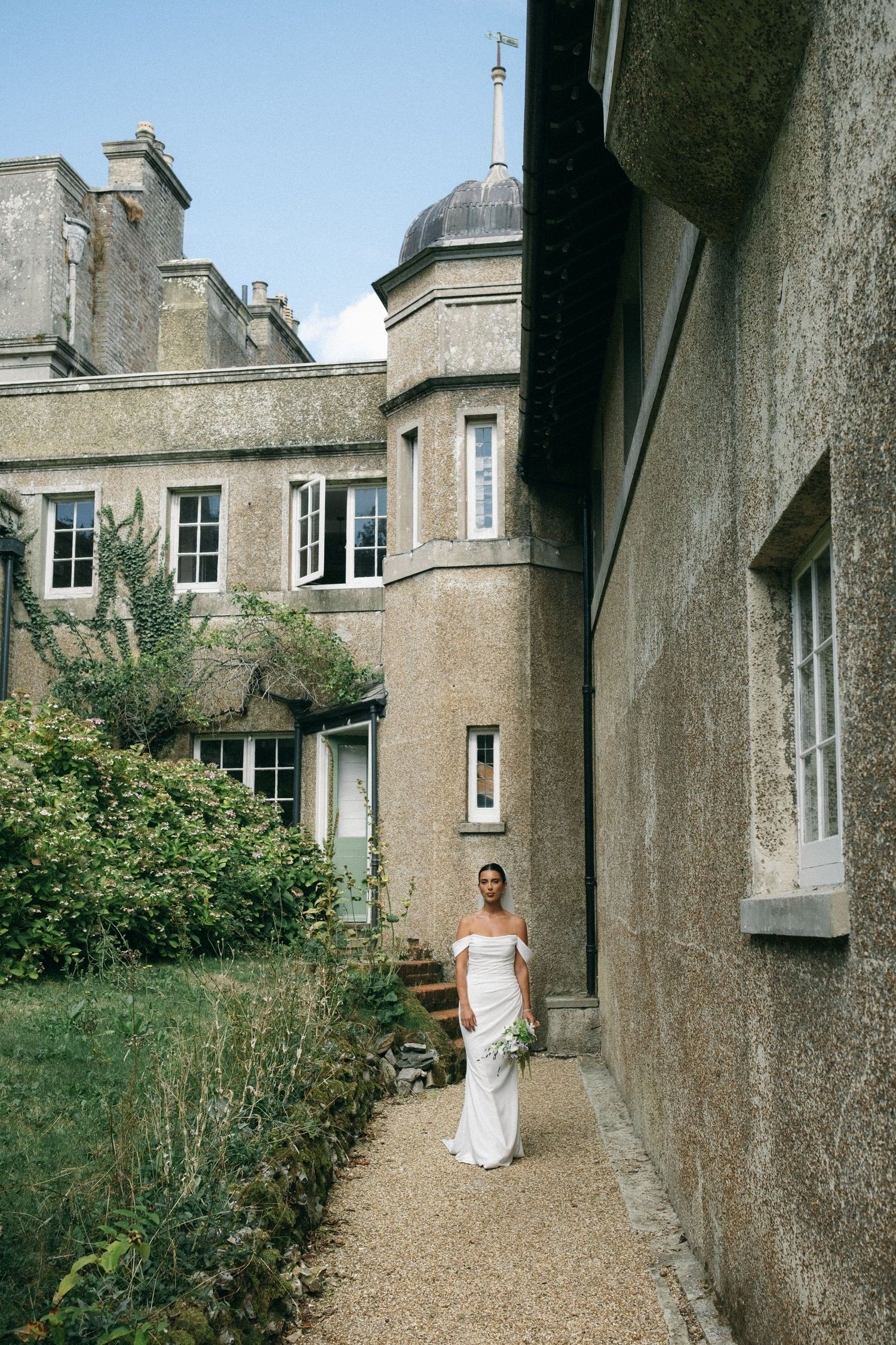 Bride in white off-shoulder gown holding bouquet stands on gravel path beside elegant stone manor house.