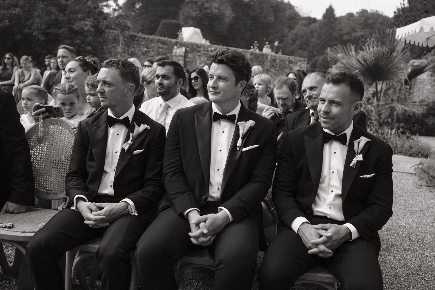 Black and white photo of three groomsmen in tuxedos seated at an outdoor wedding ceremony.