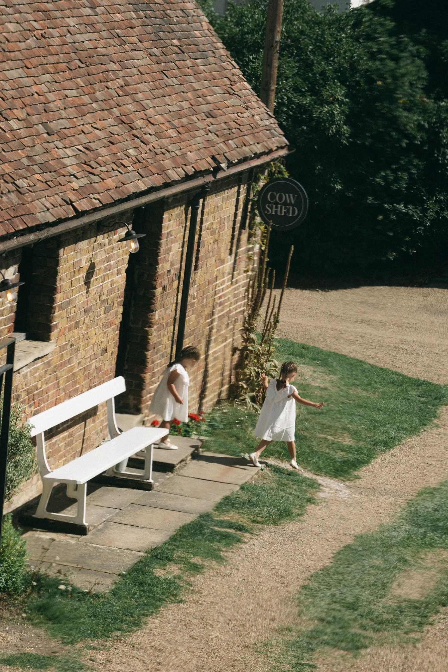Two girls in white dresses play outside a rustic brick Cow Shed building with a white bench nearby.
