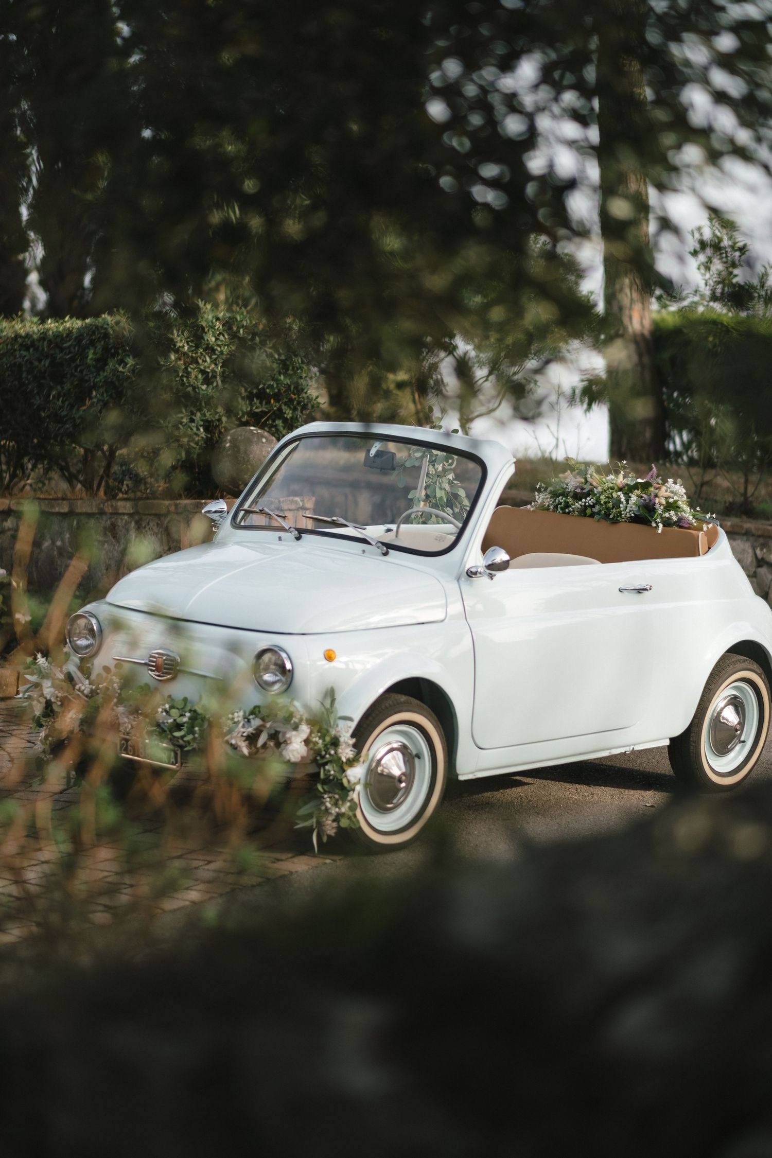 Vintage white Fiat 500 convertible decorated with flowers and greenery parked among lush green trees.