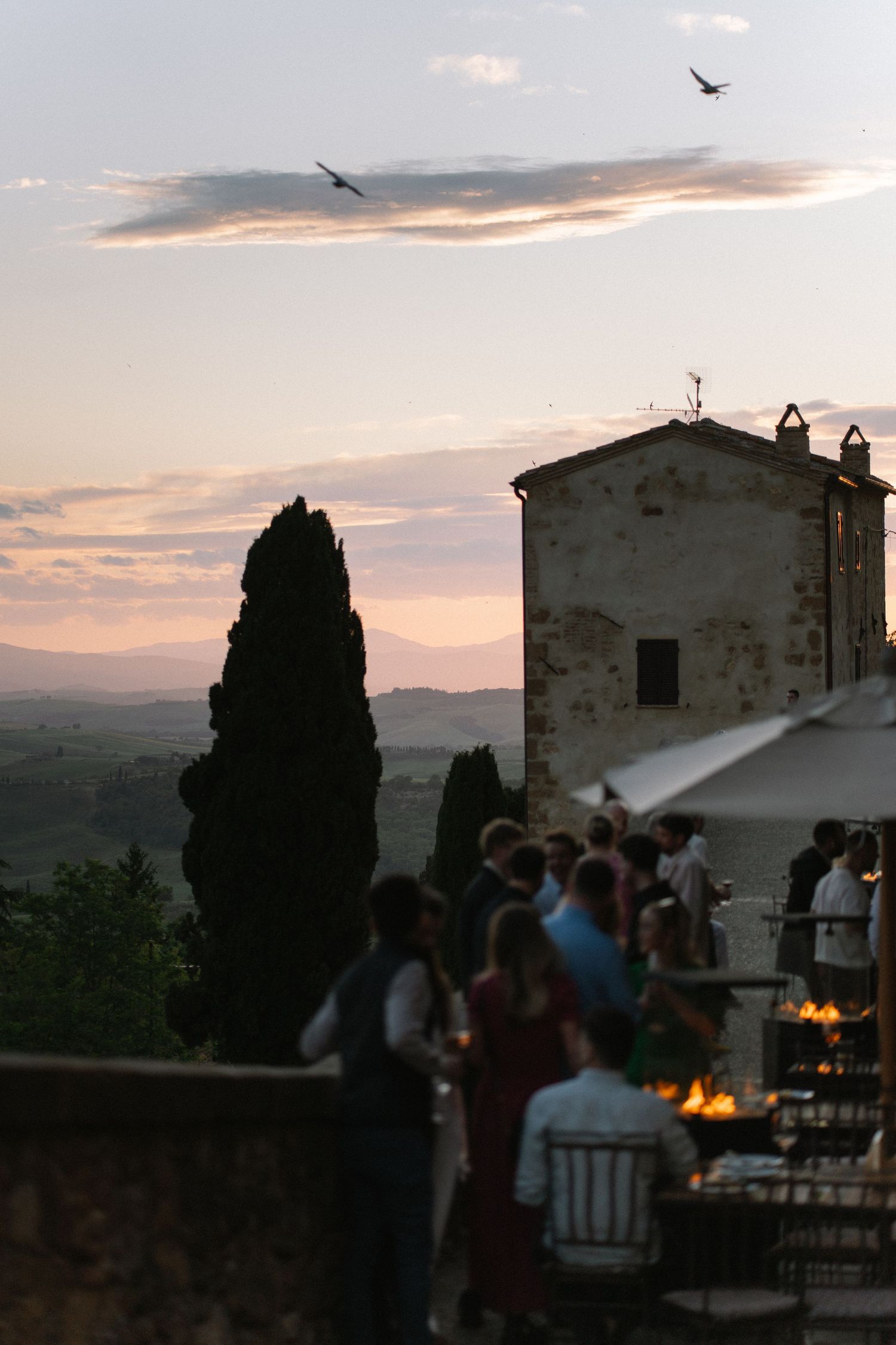 Wedding guests mingle at sunset on a Tuscan villa terrace with cypress trees and rolling hills in the background.