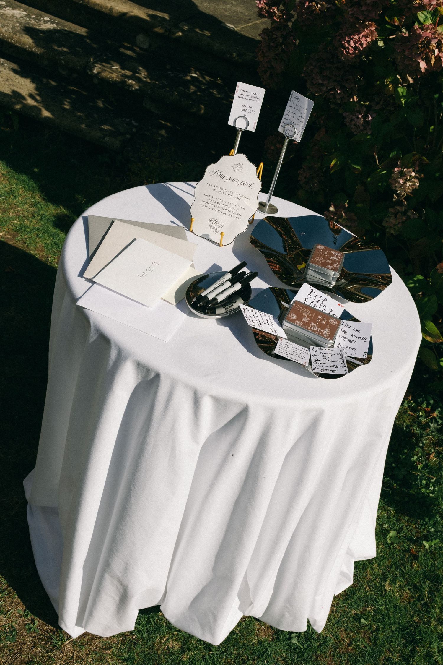 Elegant outdoor cocktail table with white linen cloth displaying wedding stationery and chocolates in garden setting.