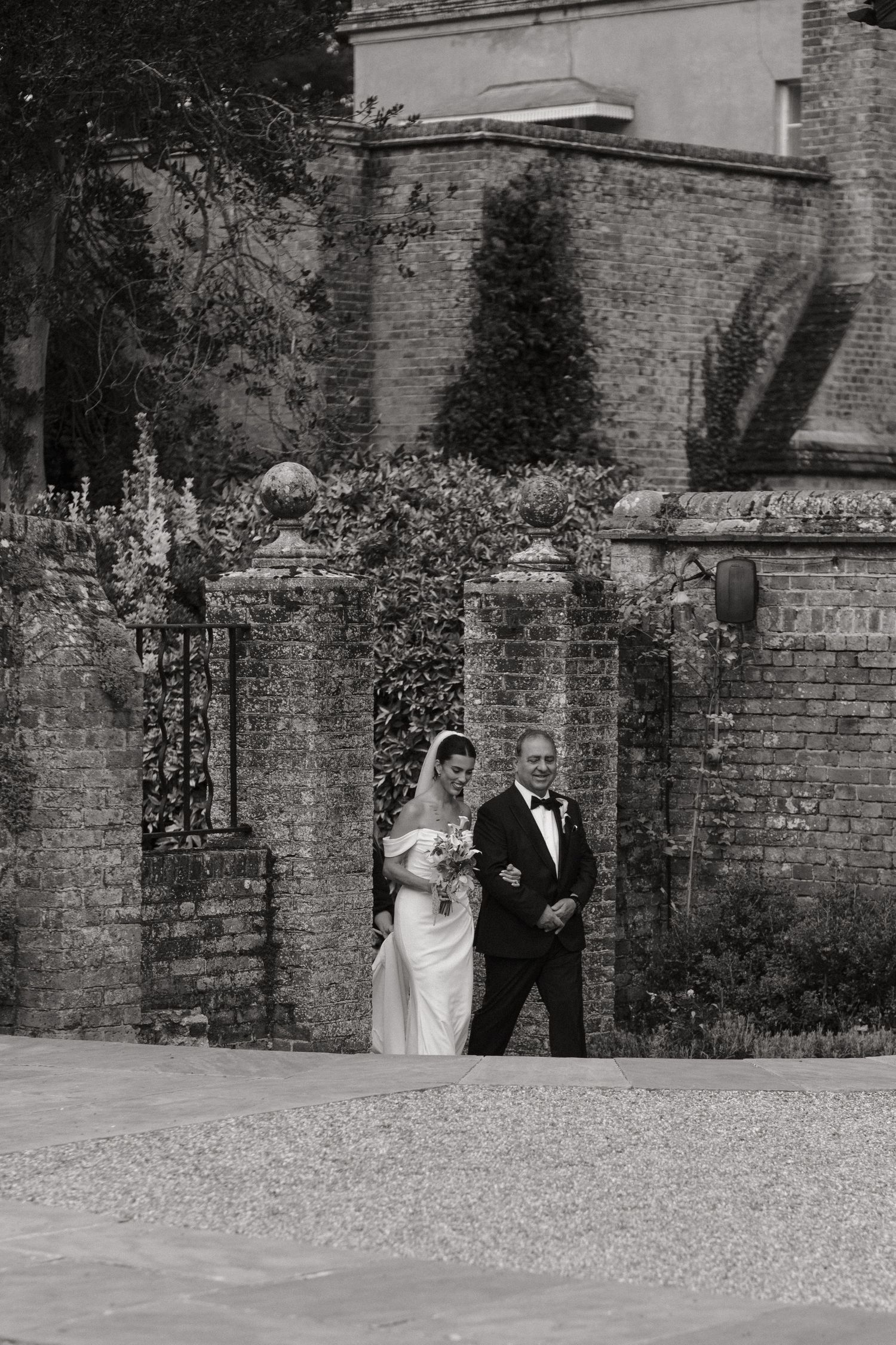 Black and white photo of a bride and groom walking through ornate stone gates at a classic English countryside wedding venue.