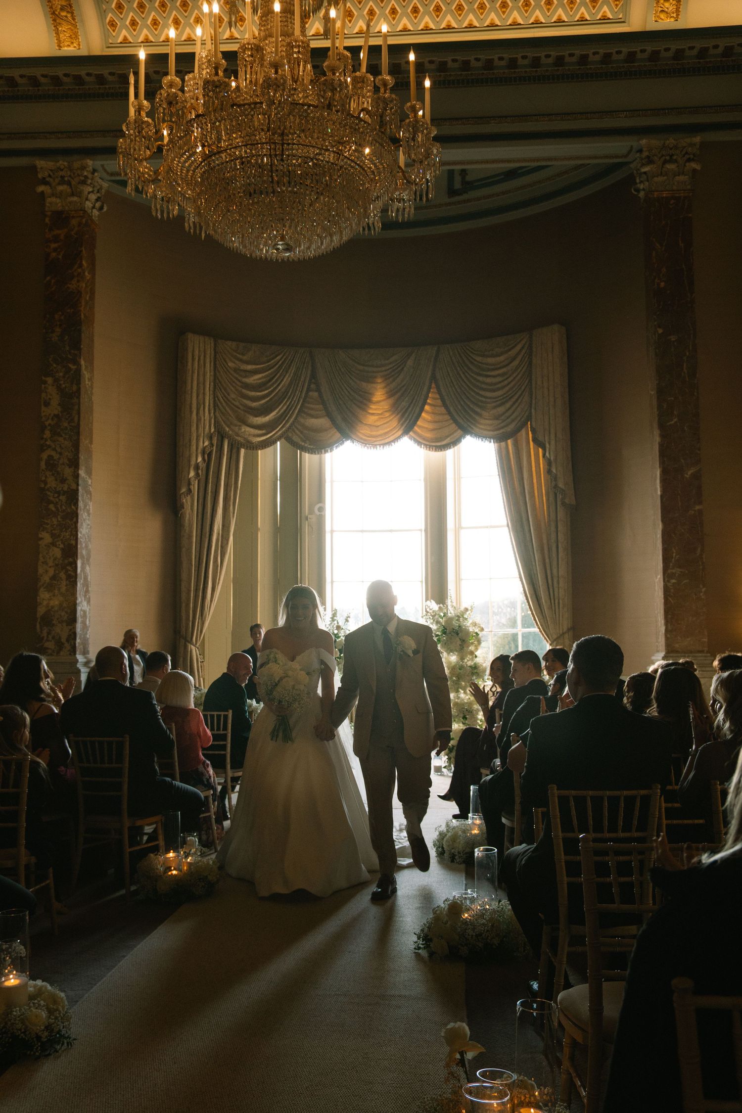 Bride and groom walk down the aisle in an elegant ballroom with chandelier and candlelit guests.