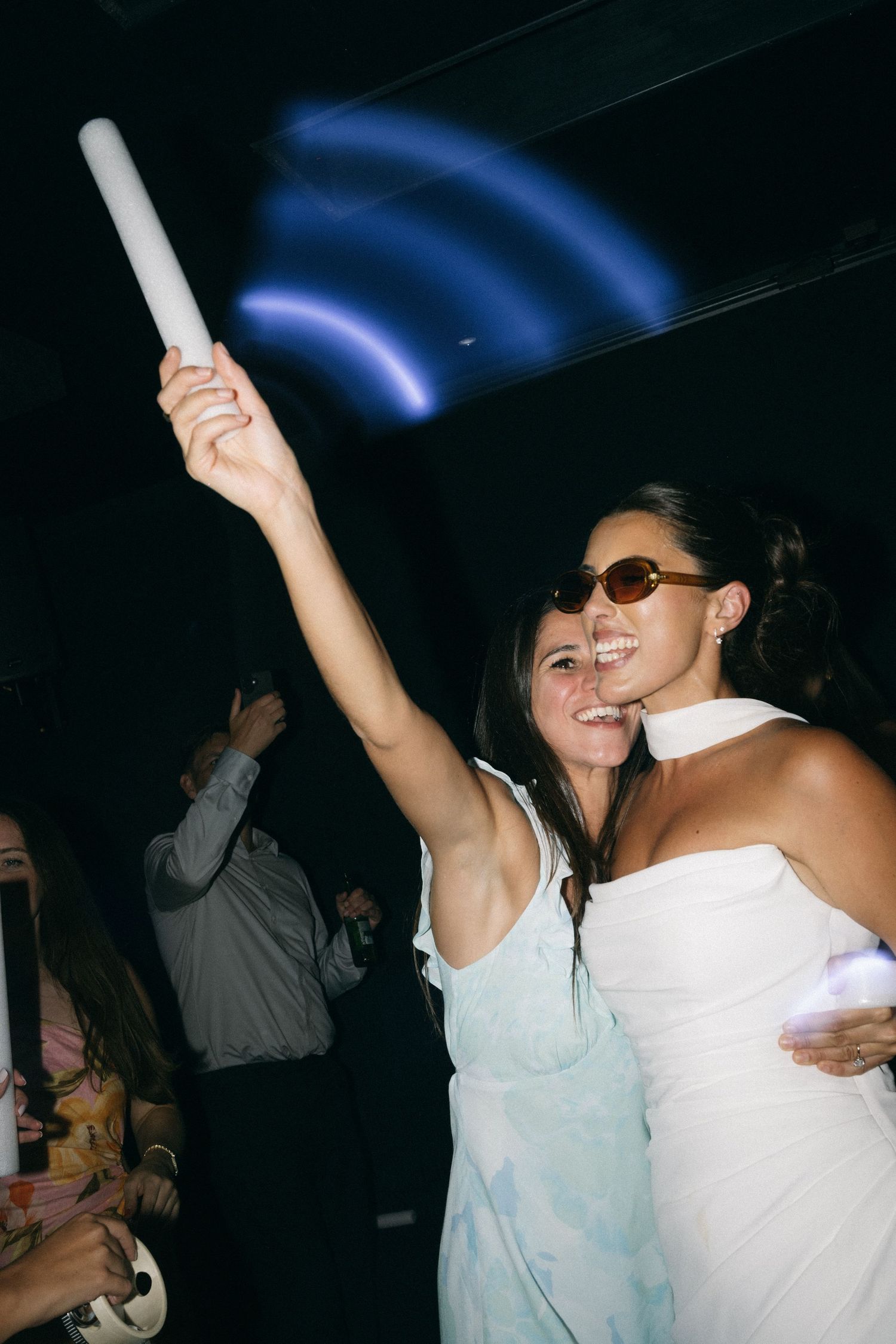 Two women dancing at a party, one holding a glow stick up, wearing white outfits and sunglasses.