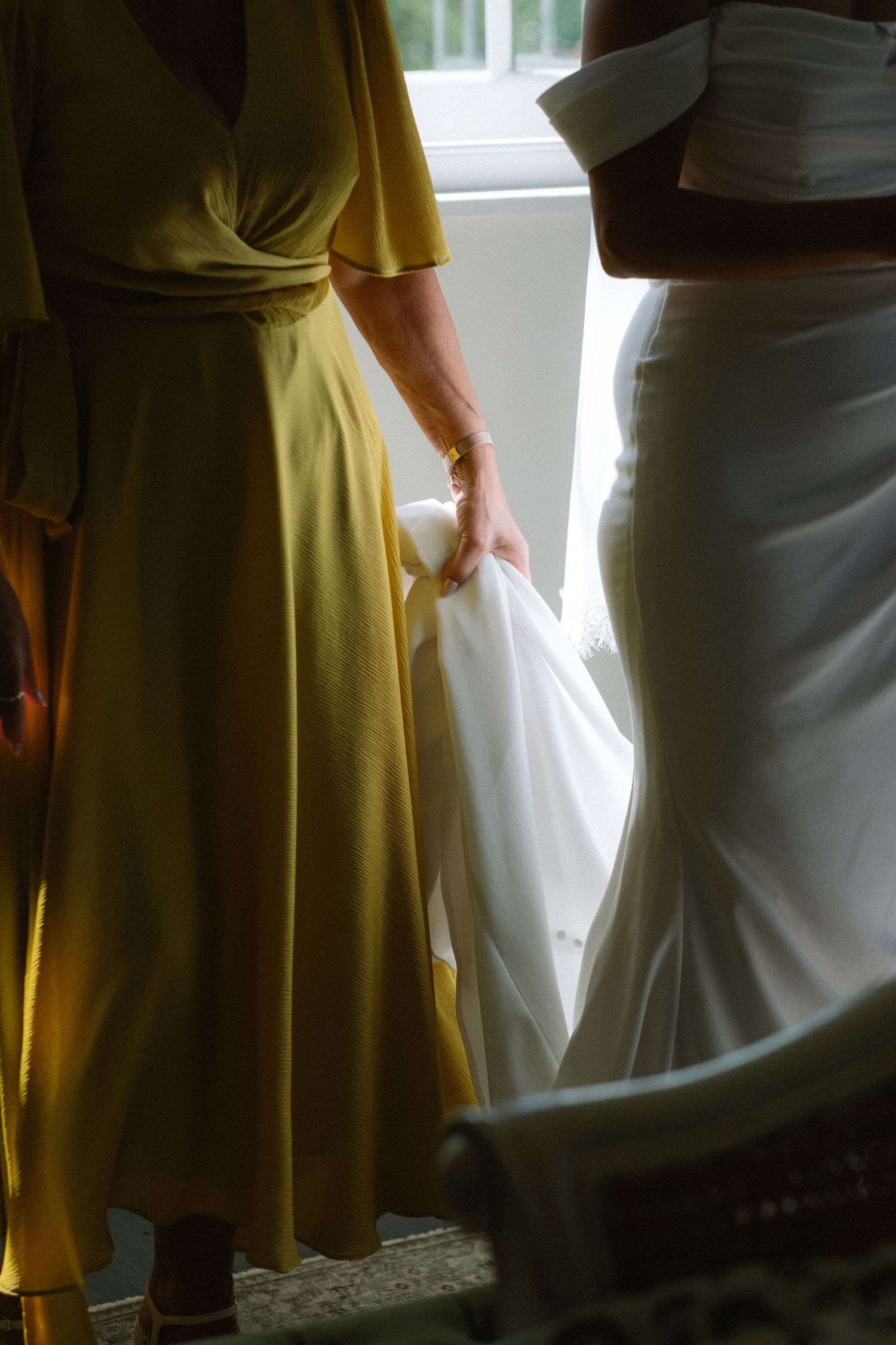 Bridesmaid in yellow dress helping bride with white wedding gown train near window.