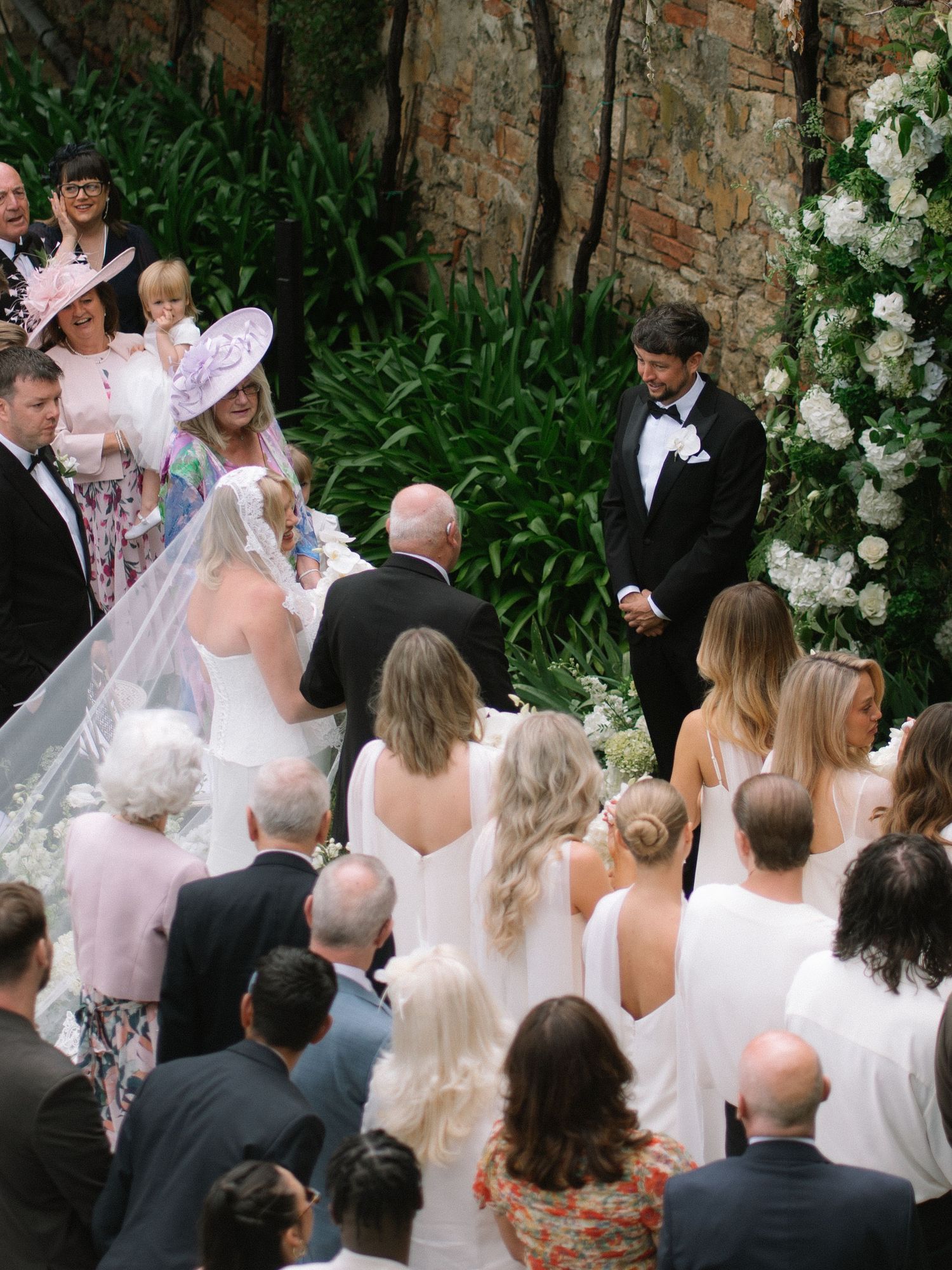 Bride in white veil arrives at outdoor wedding ceremony altar as groom in tuxedo awaits among floral arch decor.