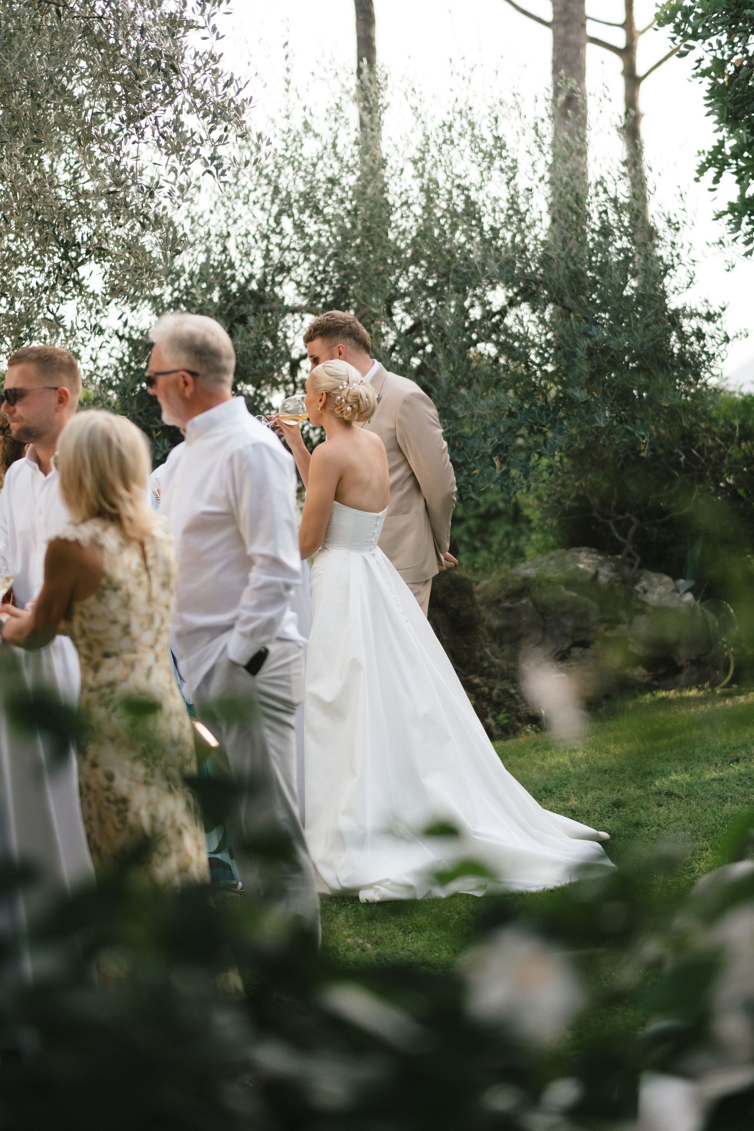Bride in flowing white gown stands with groom in tan suit during lush garden wedding ceremony.