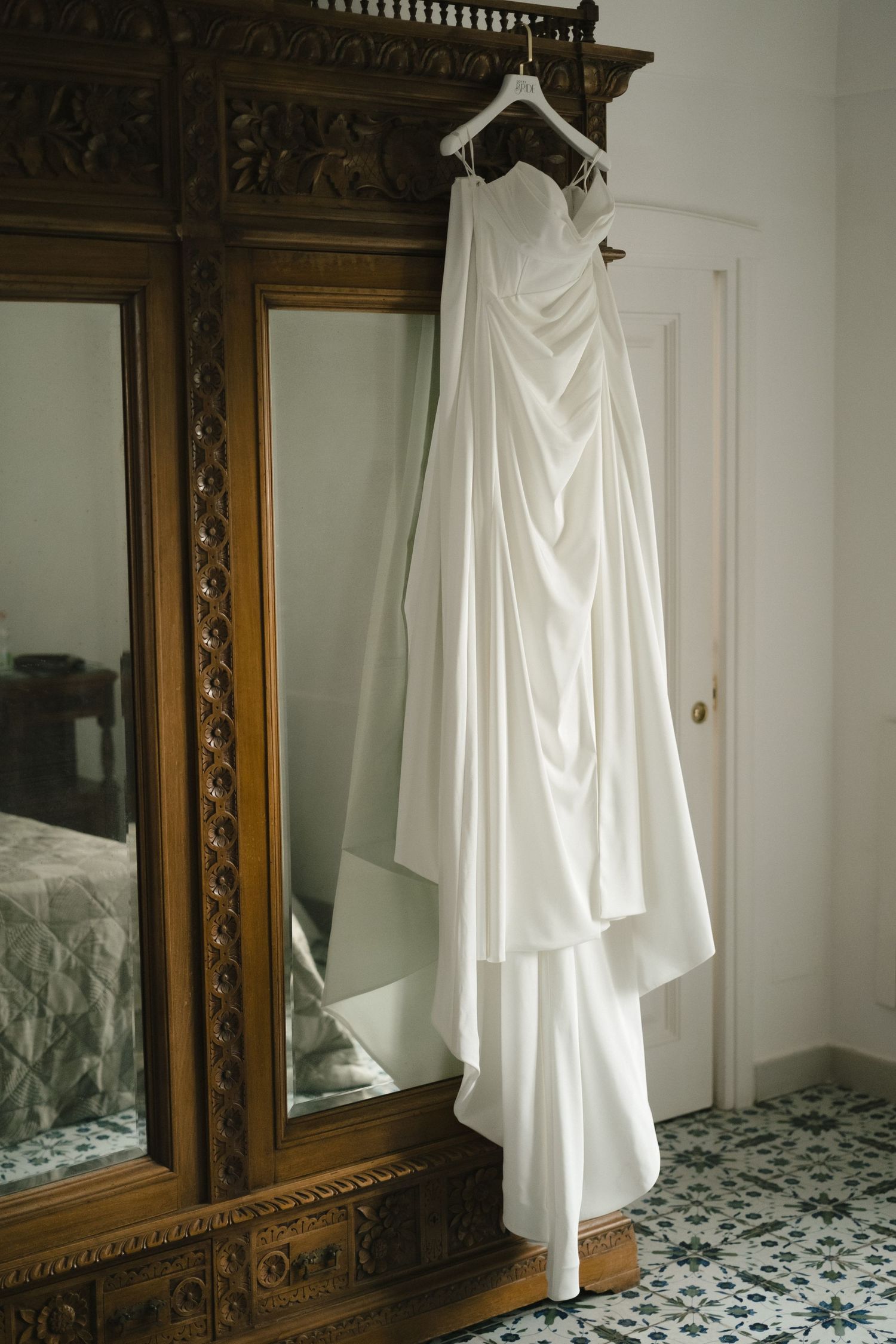 Elegant white strapless wedding gown hanging on ornate wooden armoire with mirror.