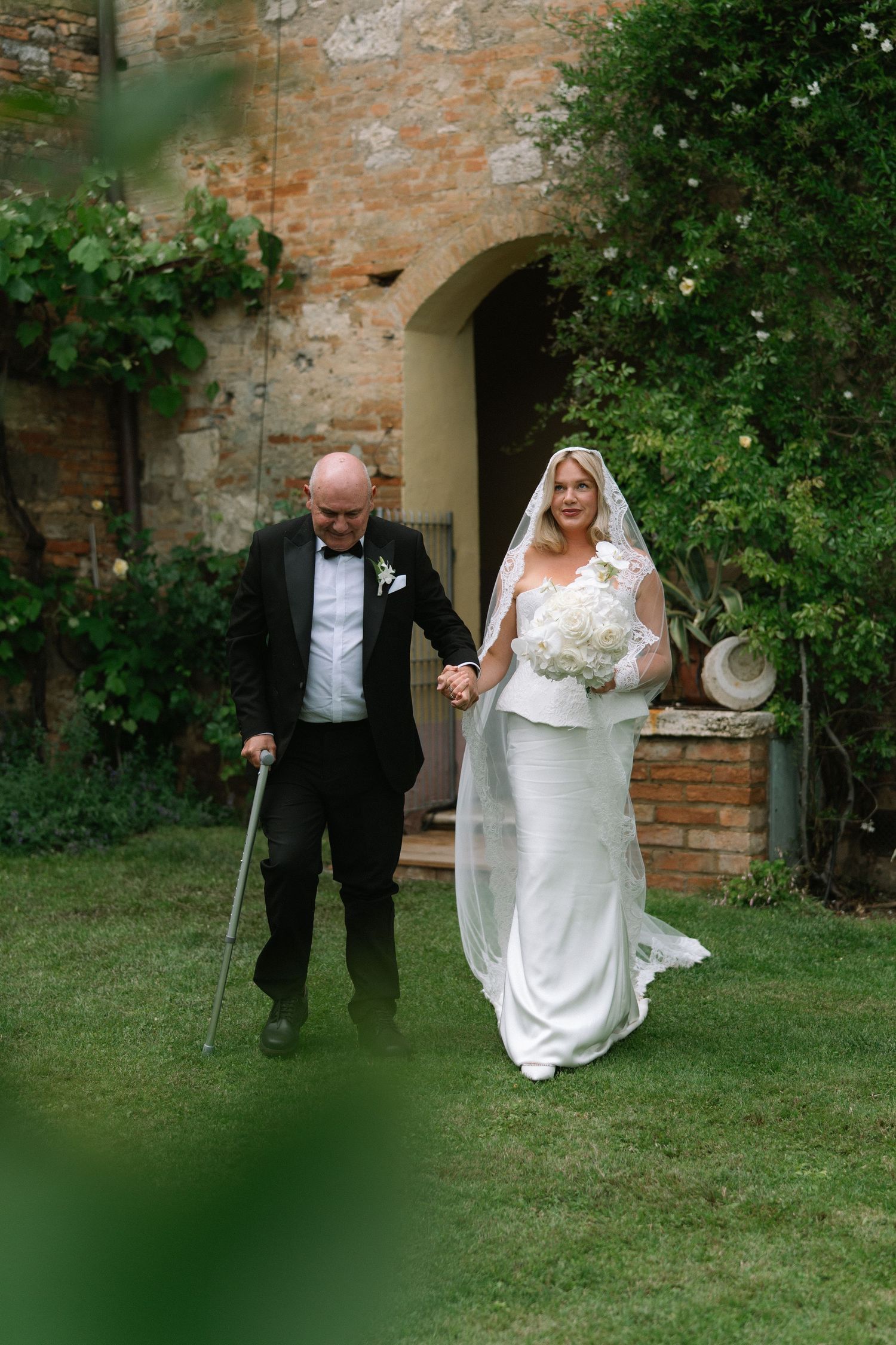 Bride in white fitted gown holding bouquet walks hand in hand with bald groom using a cane at rustic Italian venue.