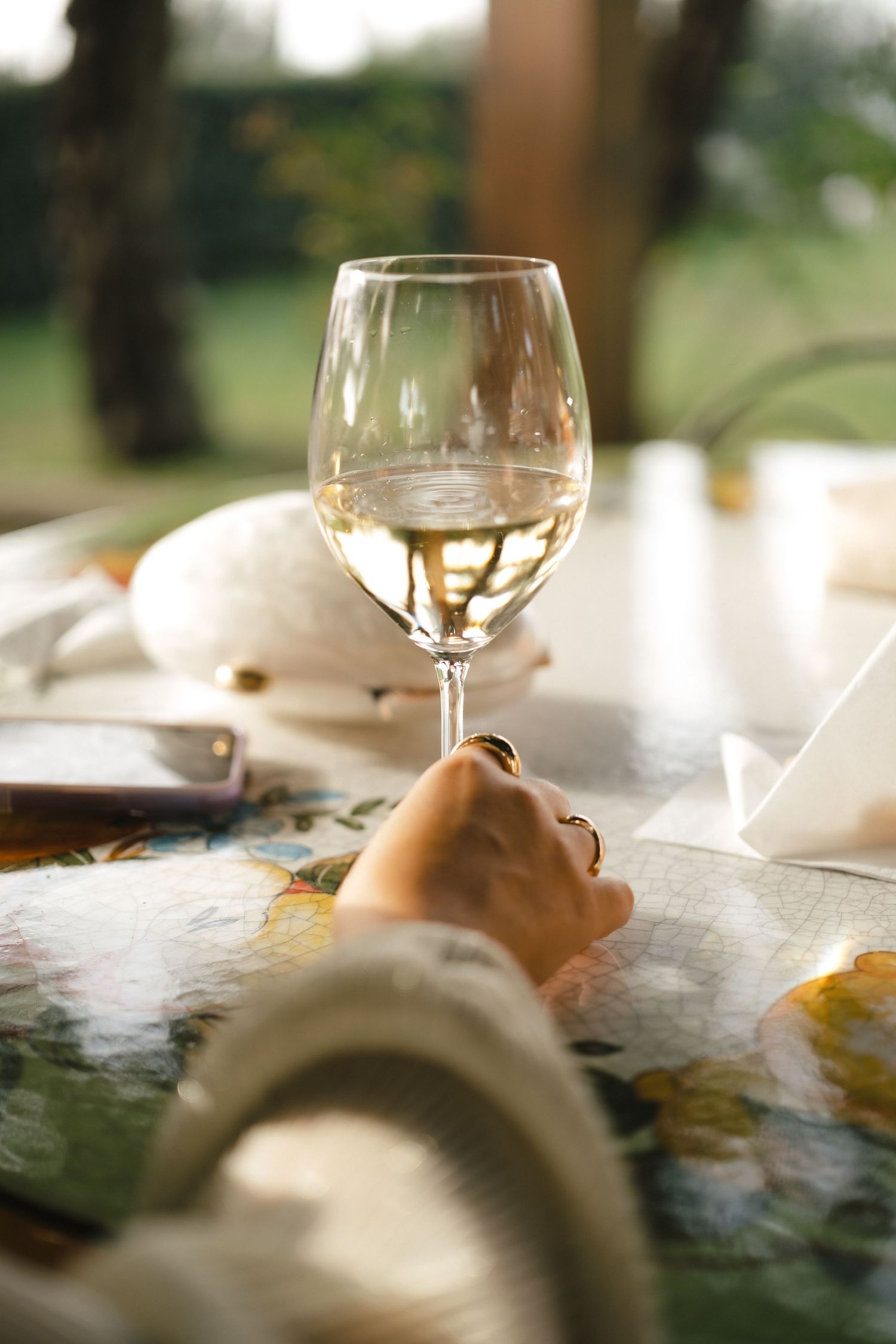 A hand resting near a glass of white wine on a floral tablecloth at an outdoor dining setting.