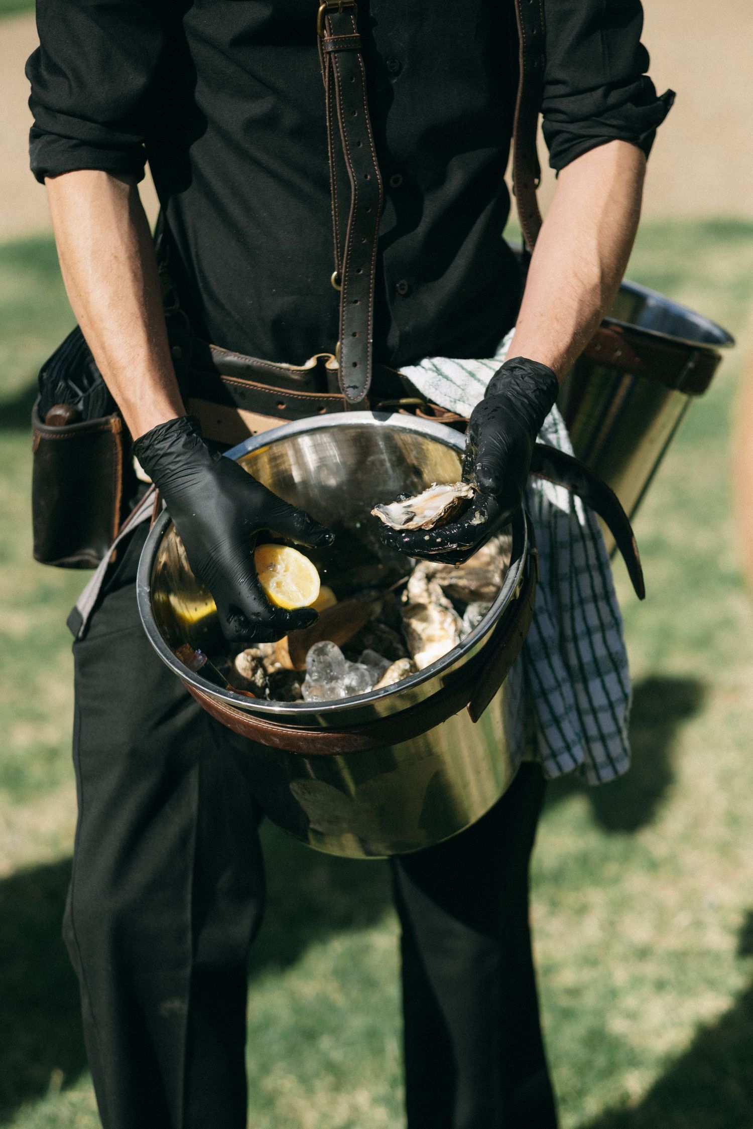 Waiter in black gloves holding a silver bucket filled with fresh oysters and ice at an outdoor catering event.