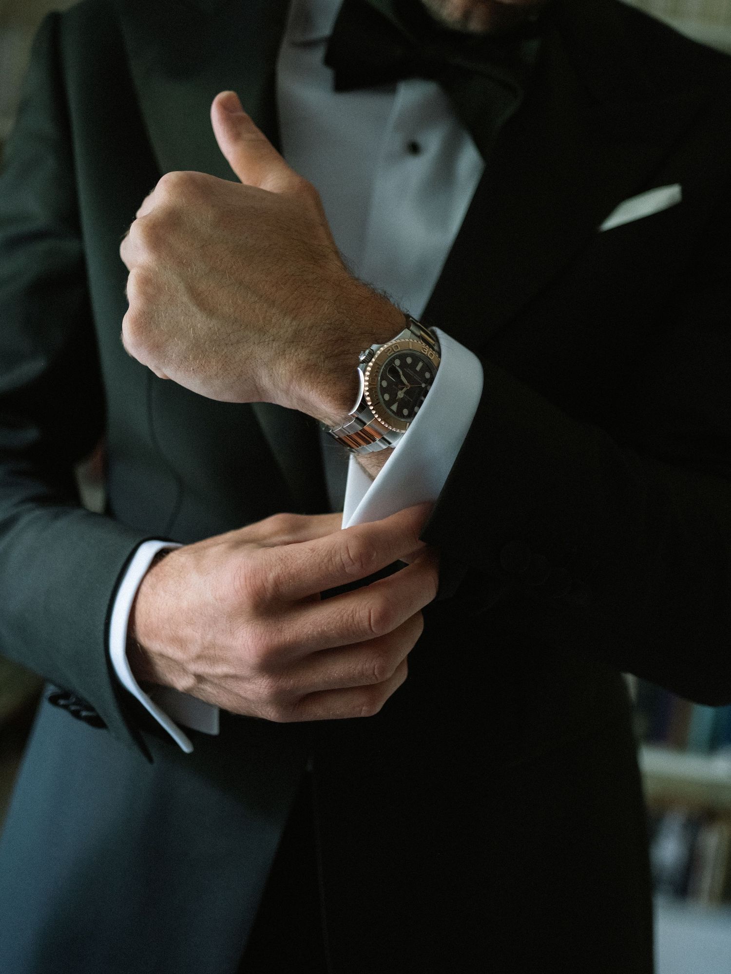 Man in black tuxedo adjusting cufflinks while wearing a luxury silver watch, dressed for a formal event.