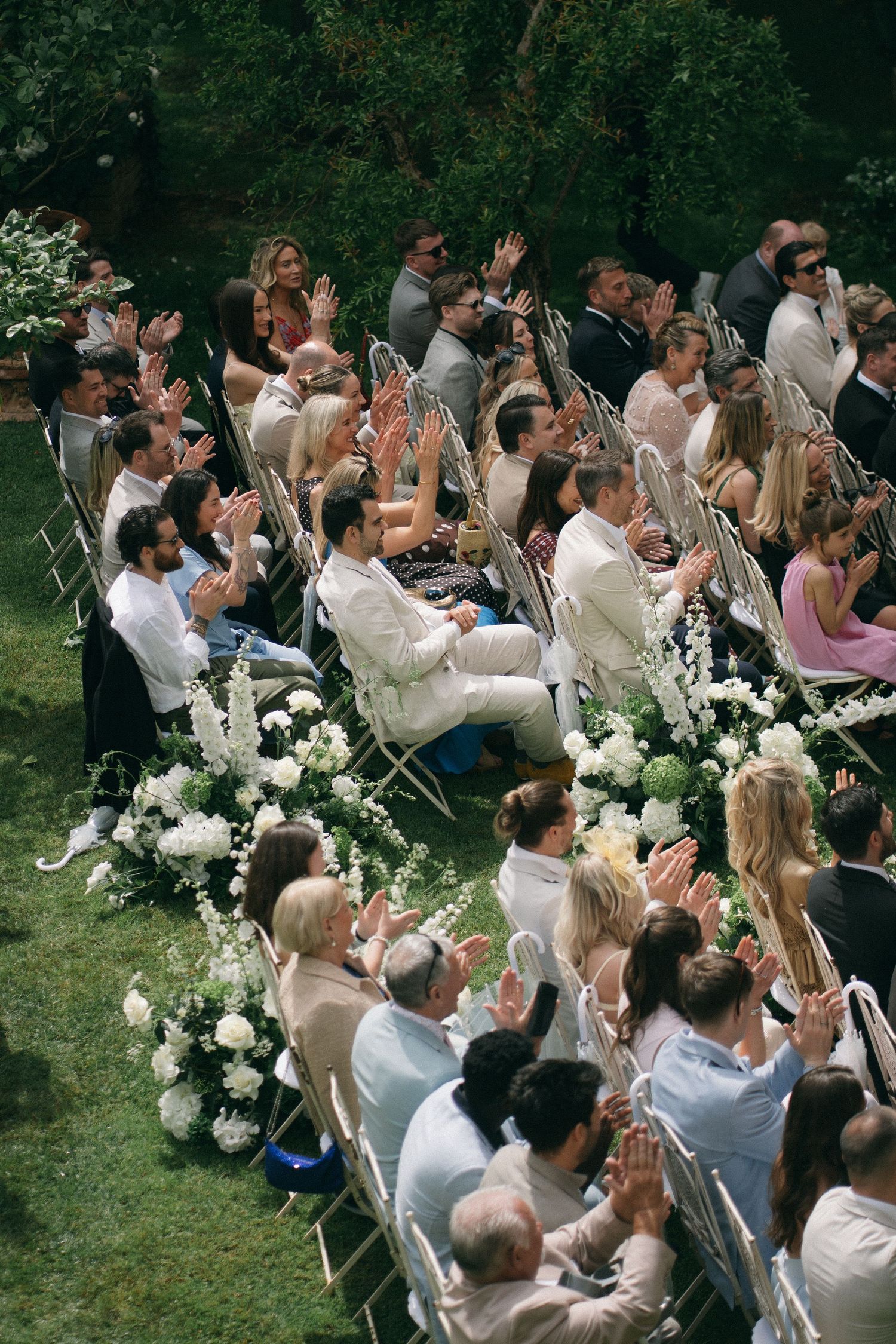 Wedding guests clap and celebrate during an outdoor garden ceremony seated on white chairs.