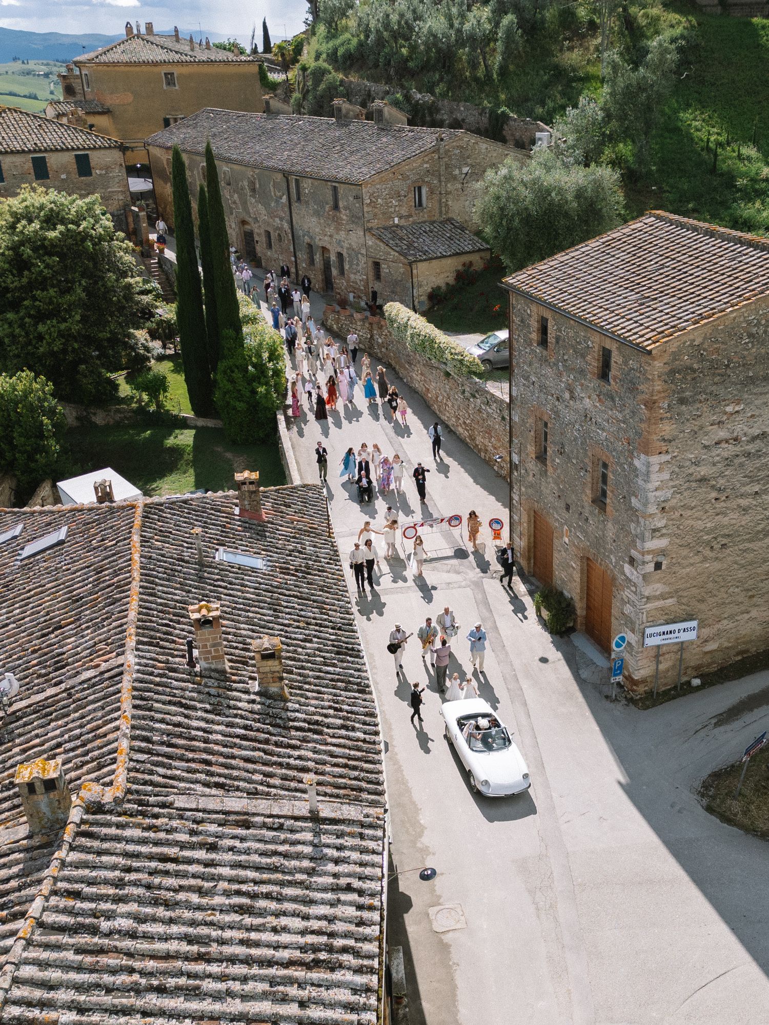 Aerial view of wedding guests and a white convertible car processing down a narrow Tuscan village road.