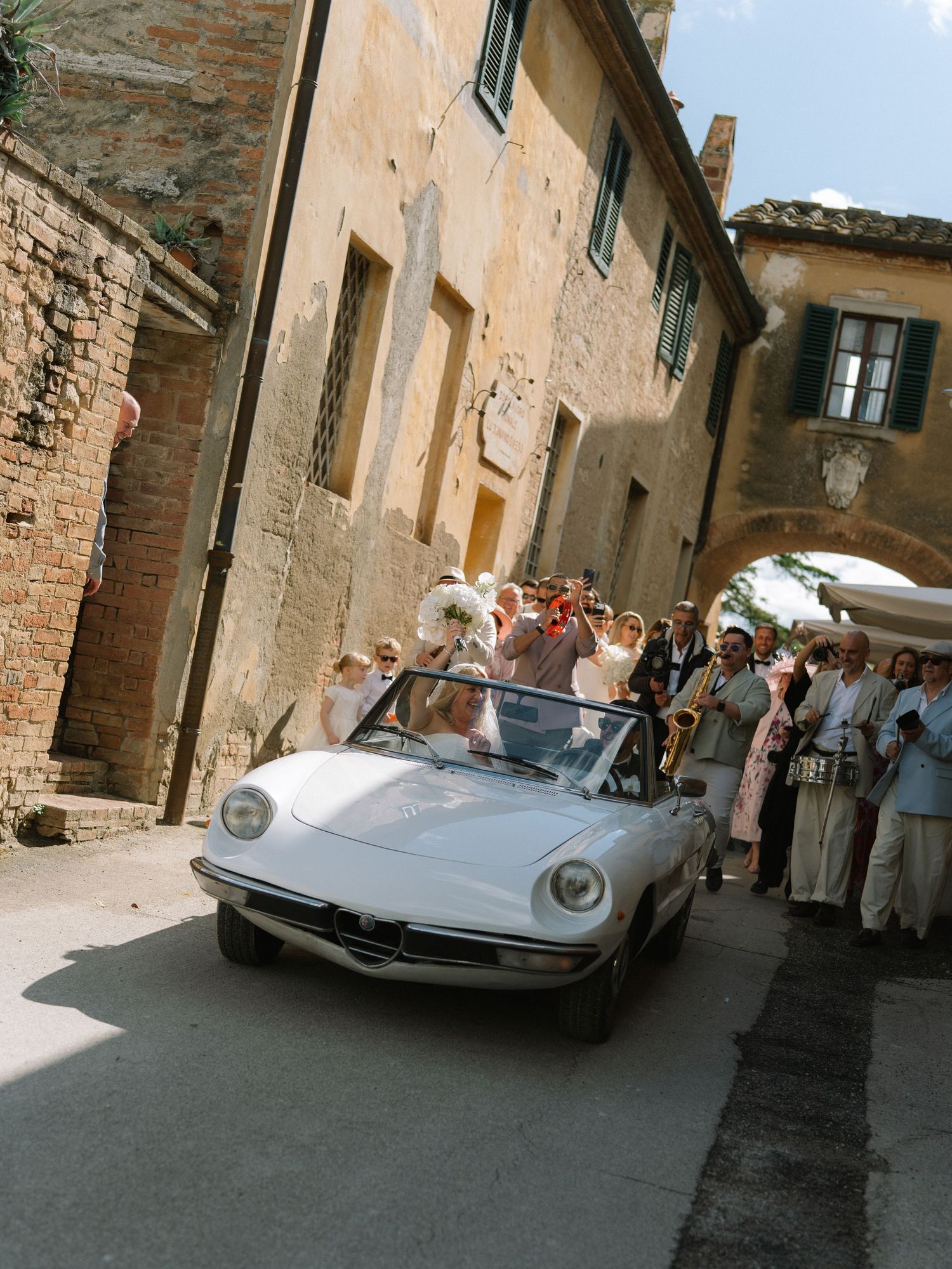 Bride and groom riding in vintage white convertible through charming Tuscan village streets with wedding guests.
