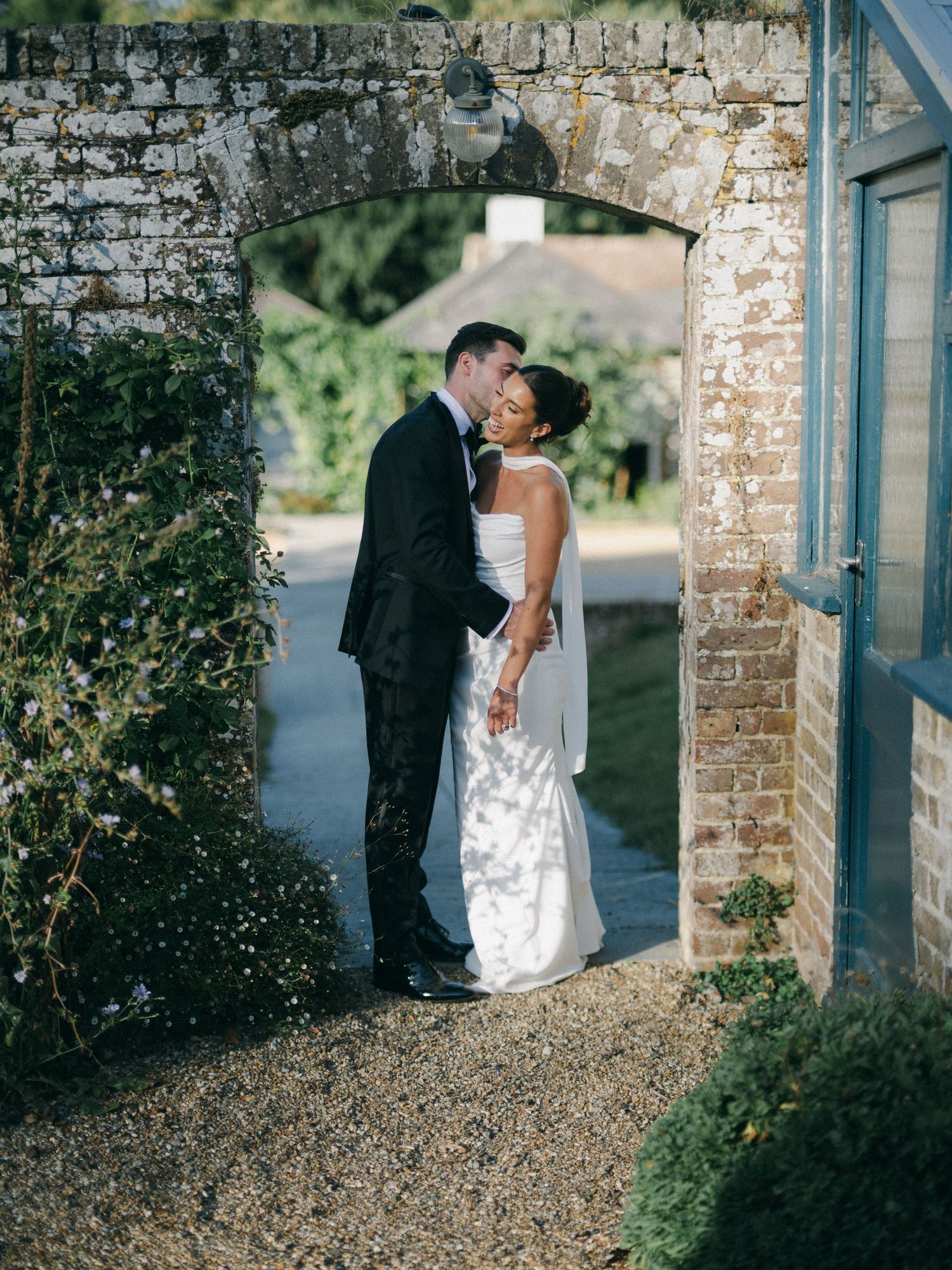 Bride and groom share a romantic kiss under a brick archway in a sunlit garden wedding venue.