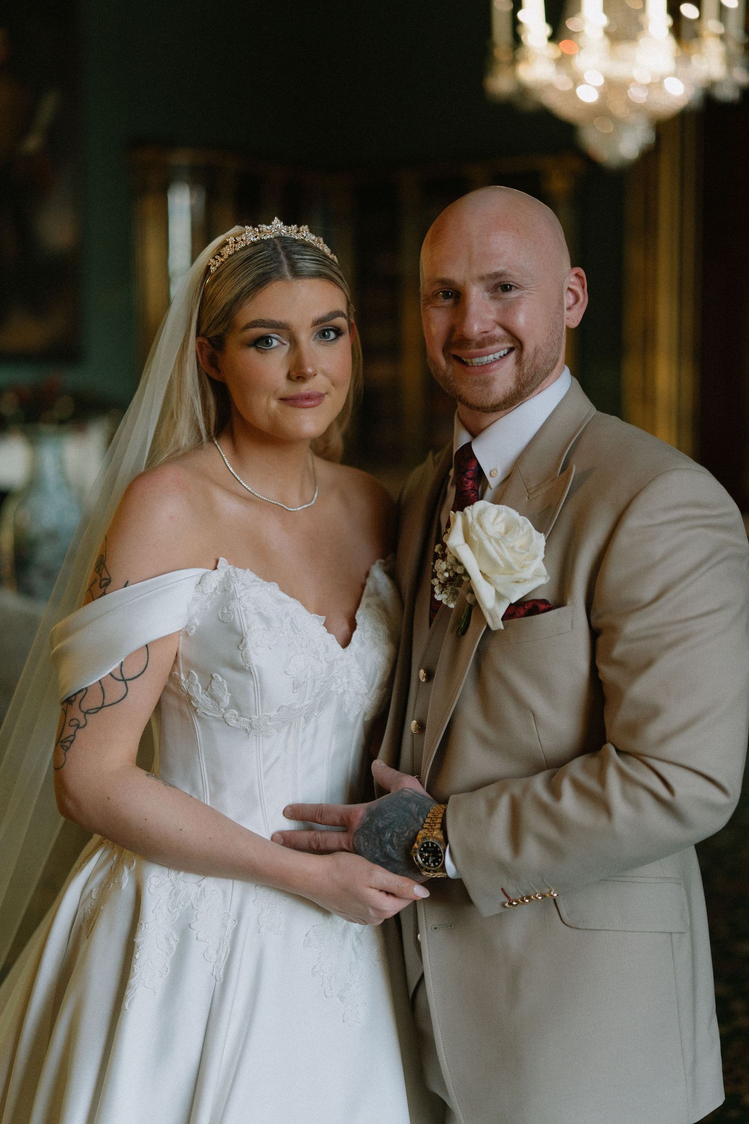 Bride in white off-shoulder gown with veil and groom in tan suit with white boutonniere pose together at elegant wedding venue.