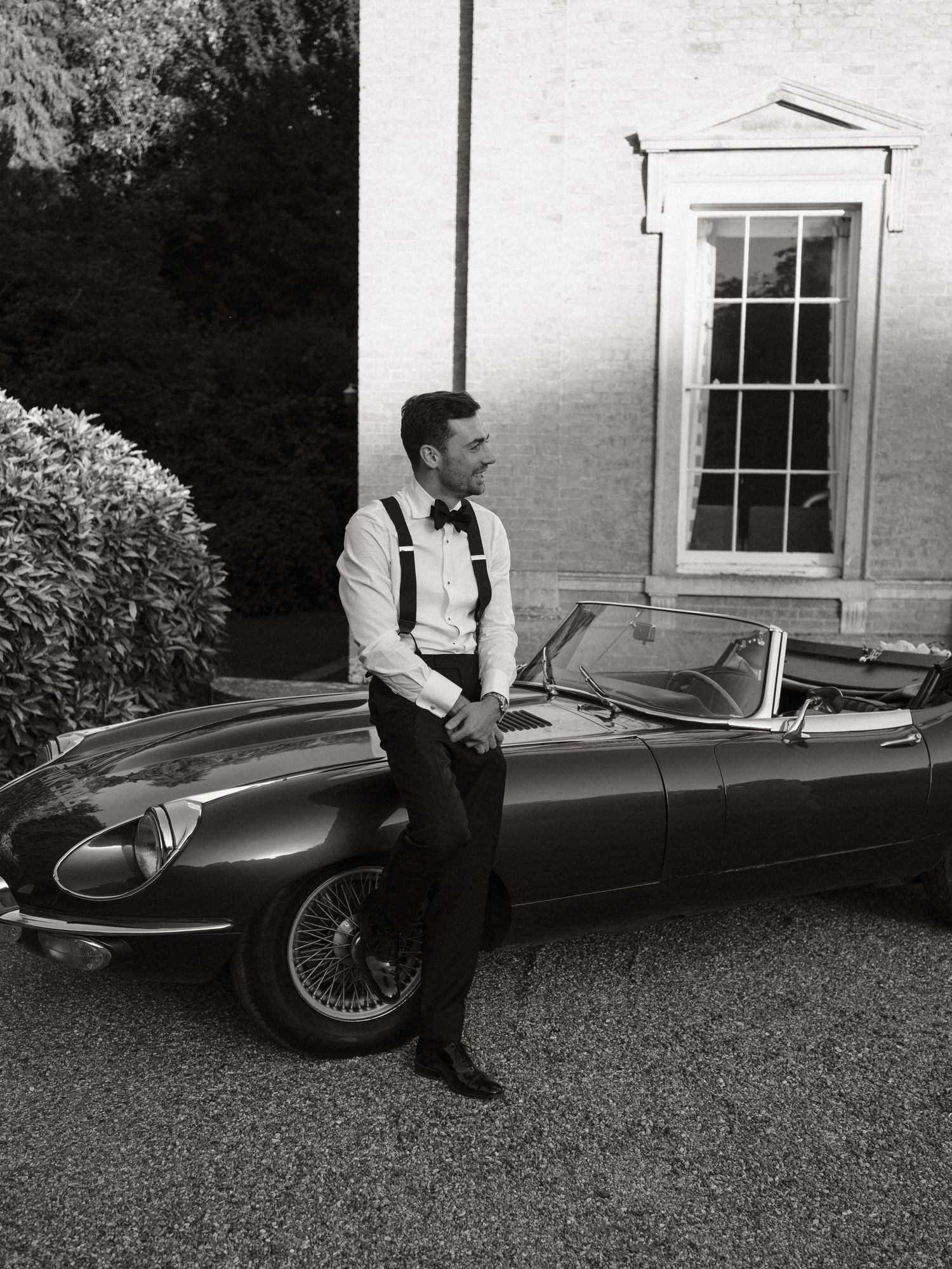Man in suspenders and bow tie leaning on a classic Jaguar E-Type convertible outside an elegant estate.