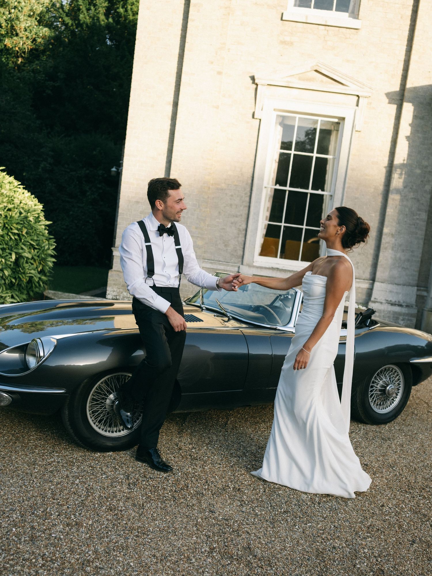 Bride and groom holding hands beside a vintage Jaguar E-Type sports car outside an elegant country house.