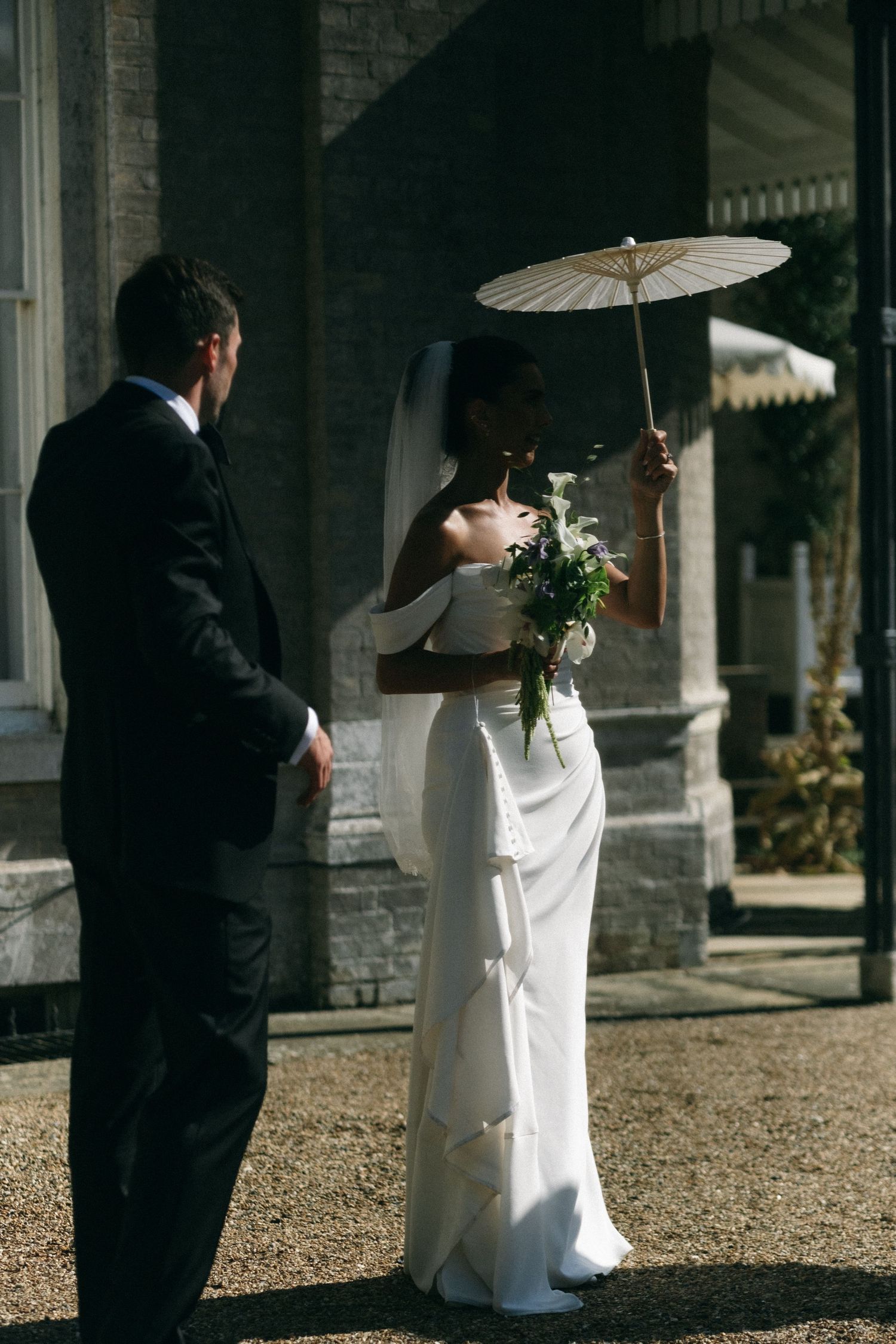 Bride in white gown holding parasol and bouquet stands with groom in black suit outside elegant stone building.
