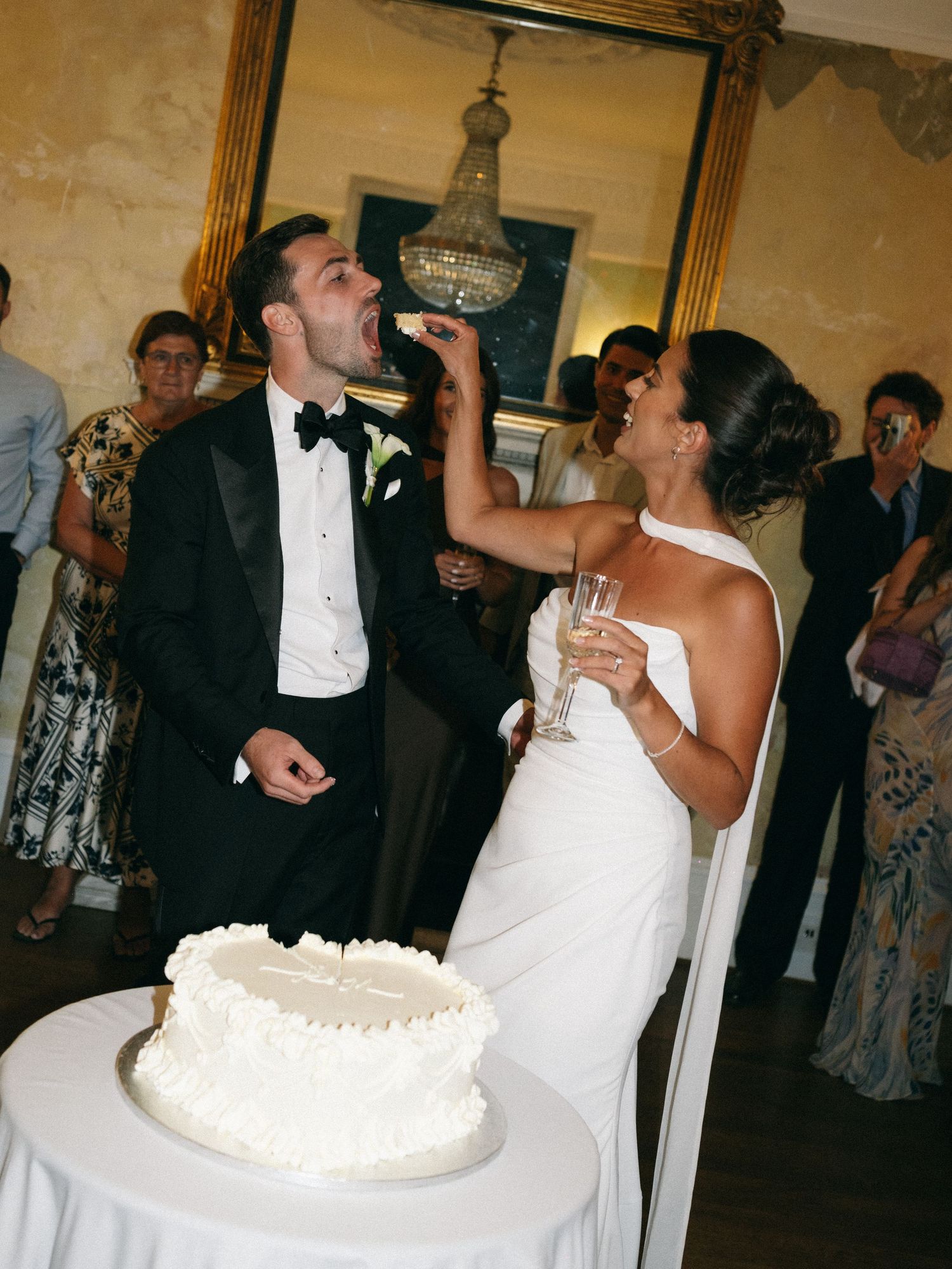 Bride feeds groom wedding cake during reception, white tiered cake on table, guests watching in elegant venue.