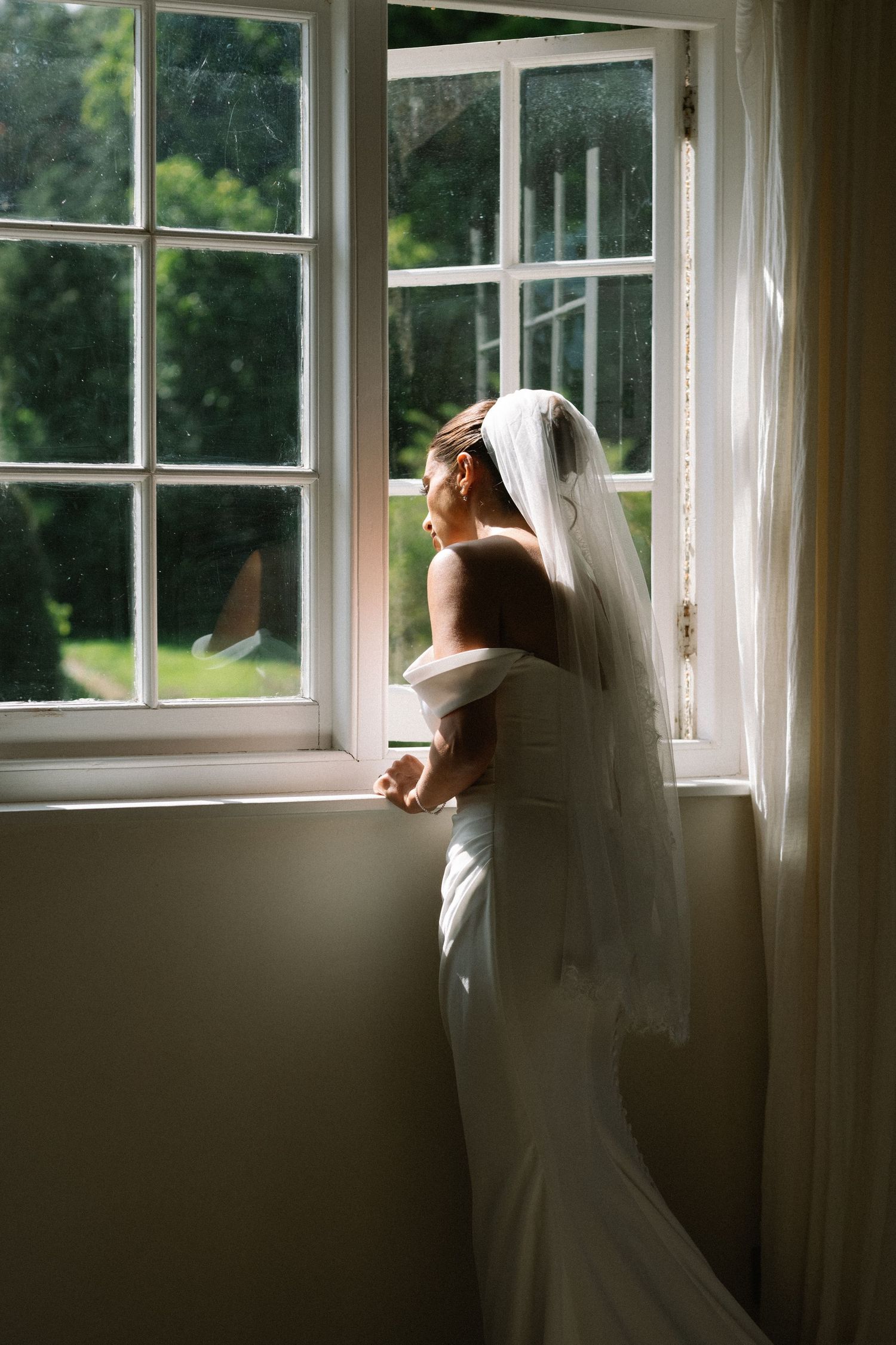 A bride in a white gown and veil stands gazing out a sunlit window on her wedding day.