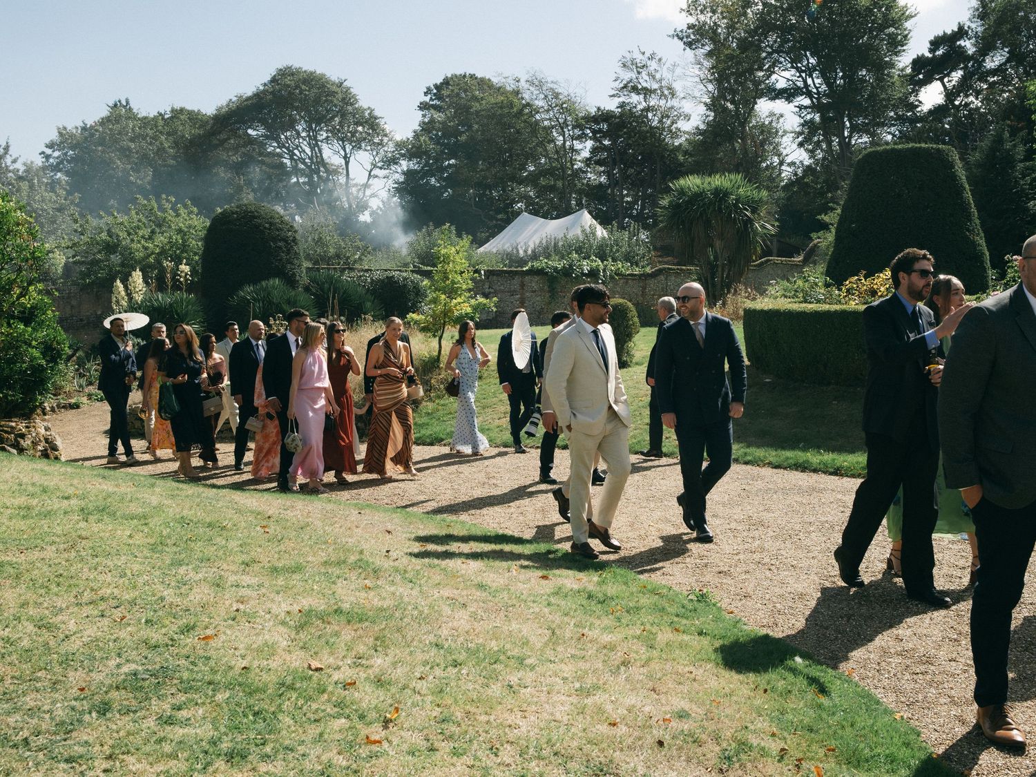 Wedding guests walking through a formal garden with topiary hedges on a sunny day.