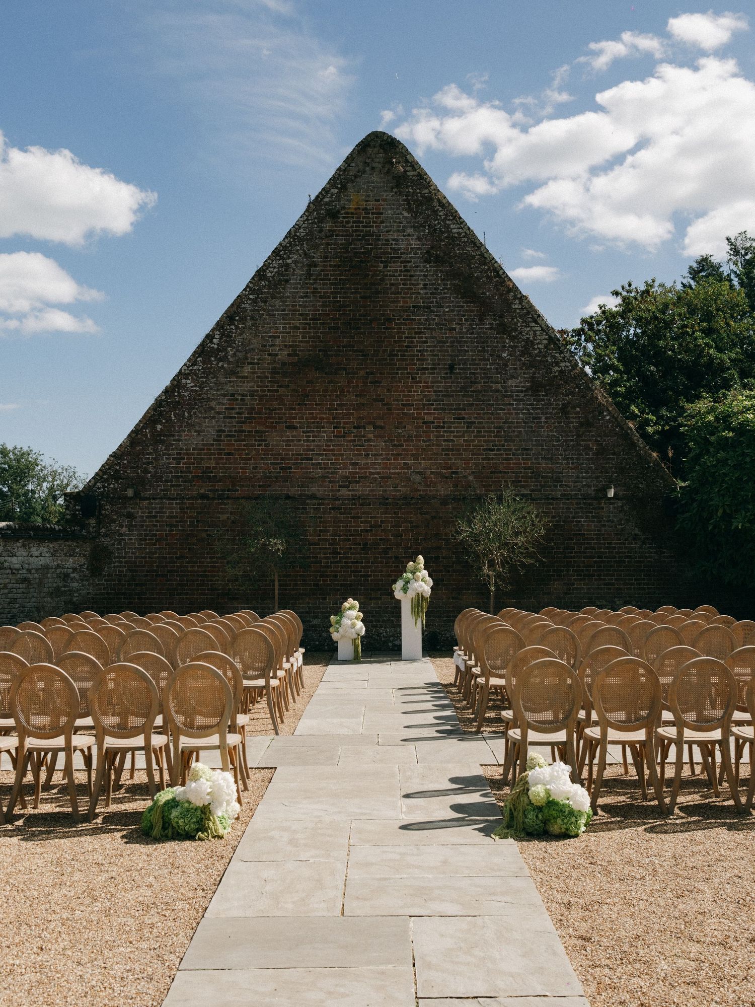 Outdoor wedding ceremony setup with wooden chairs, floral arrangements, and a rustic brick pyramid backdrop under a blue sky.