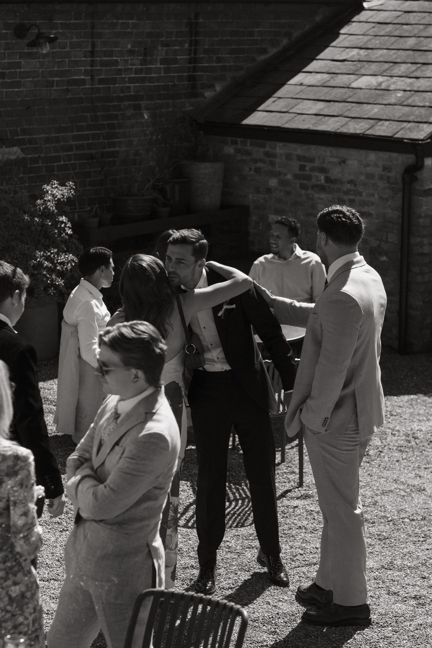 Black and white photo of elegantly dressed guests socializing outdoors at a formal event near a brick building.