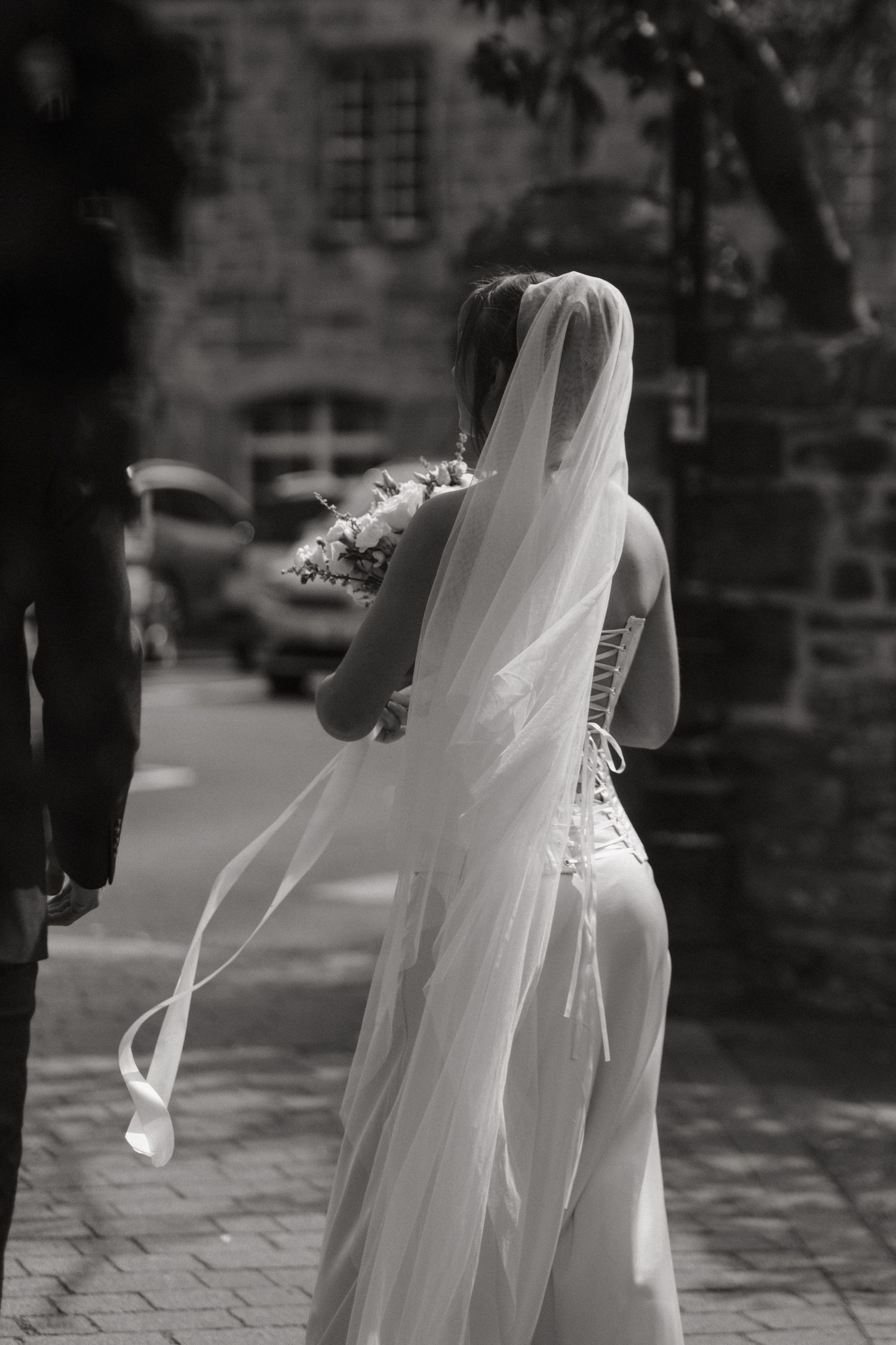 Black and white photo of a bride in a flowing veil and white gown holding a bouquet, walking along a cobblestone path.