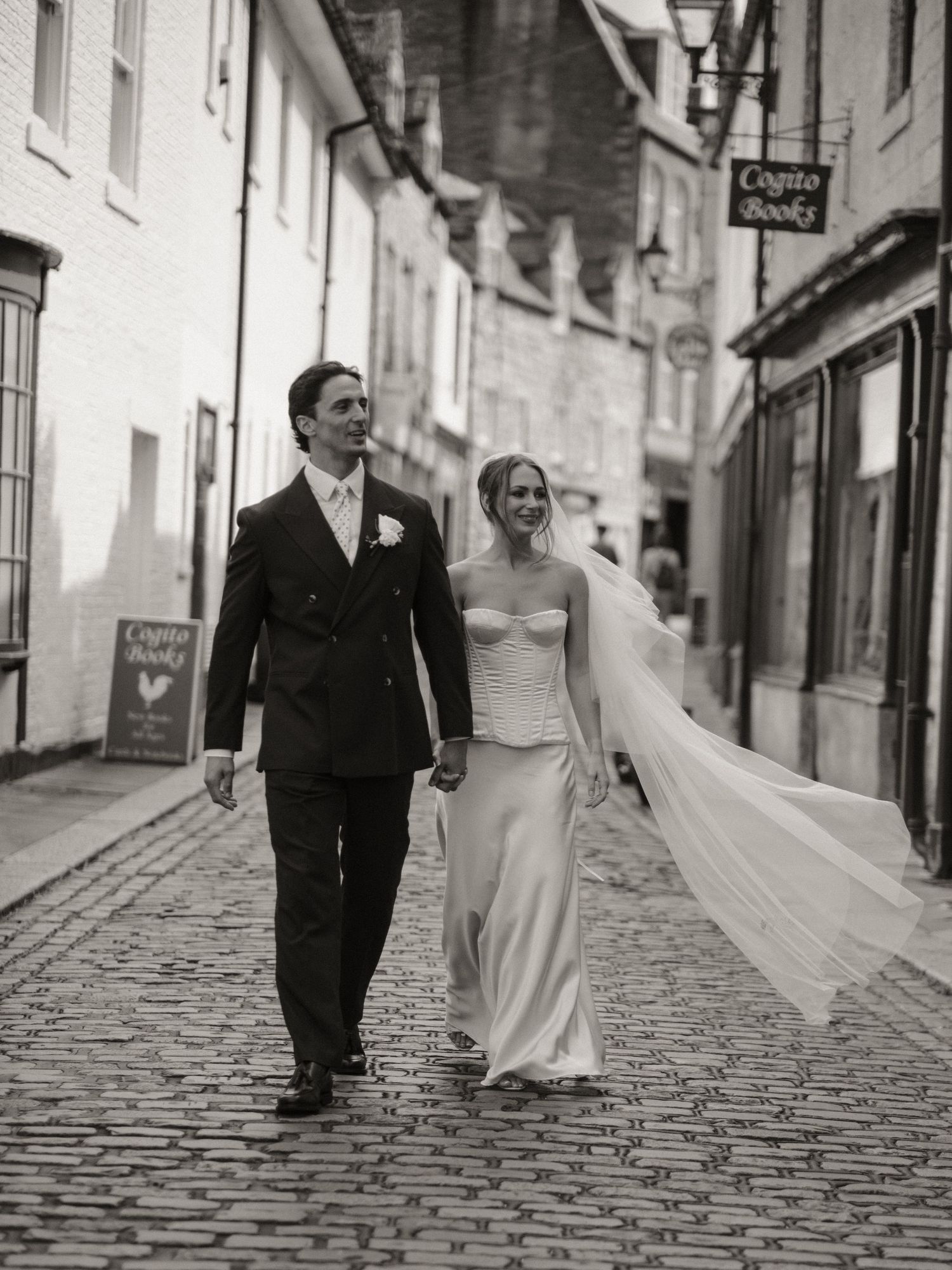 Black and white photo of bride and groom walking on a cobblestone street in a historic town.