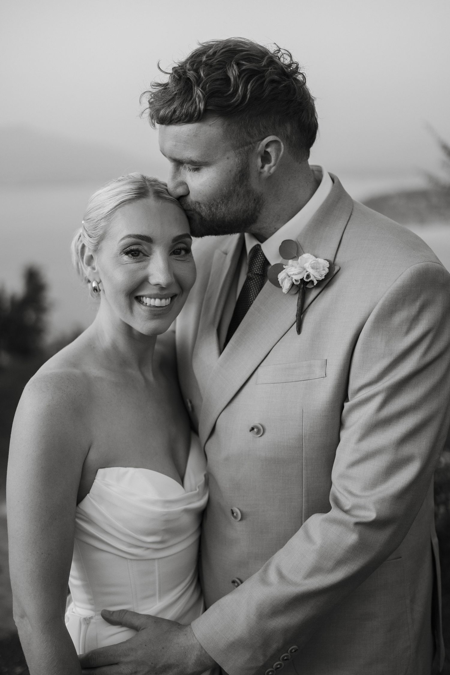 Black and white portrait of groom kissing smiling bride on forehead with scenic coastal backdrop.
